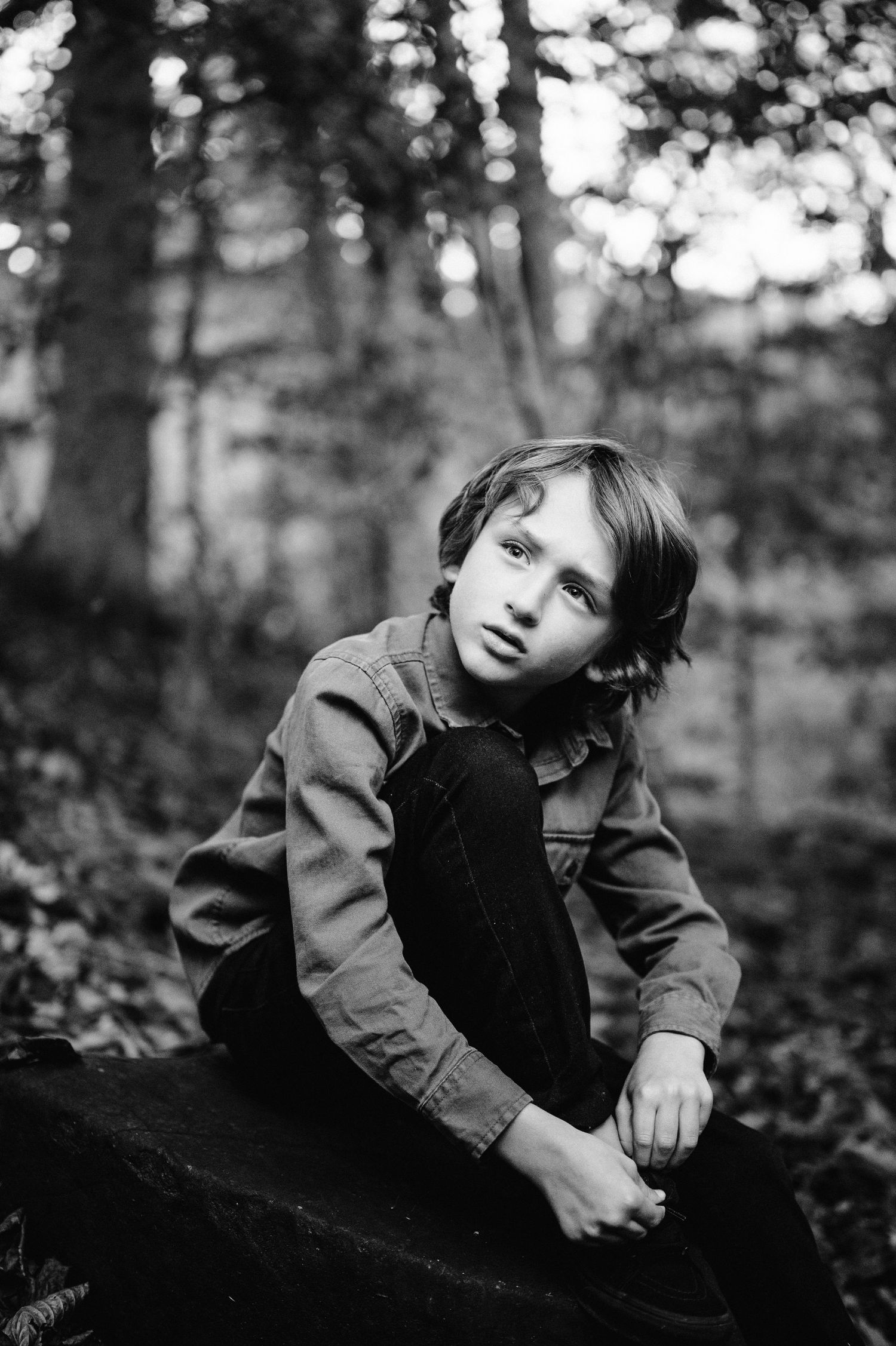 Black and white portrait of a youth sitting thoughtfully on a rock in a forest setting.