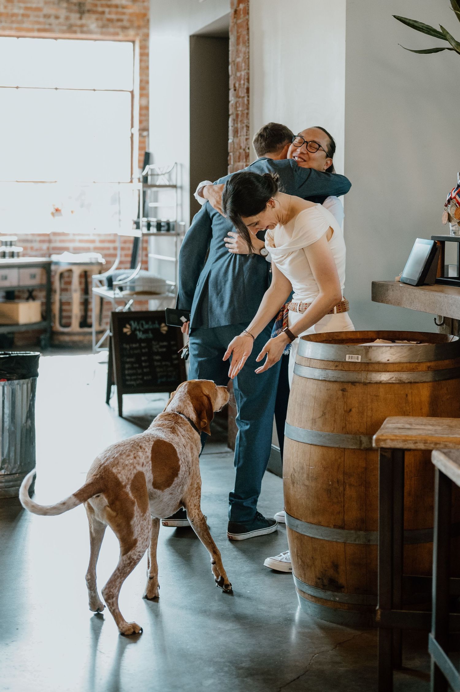 A couple embracing in a rustic venue with wooden barrels while their dog looks on.