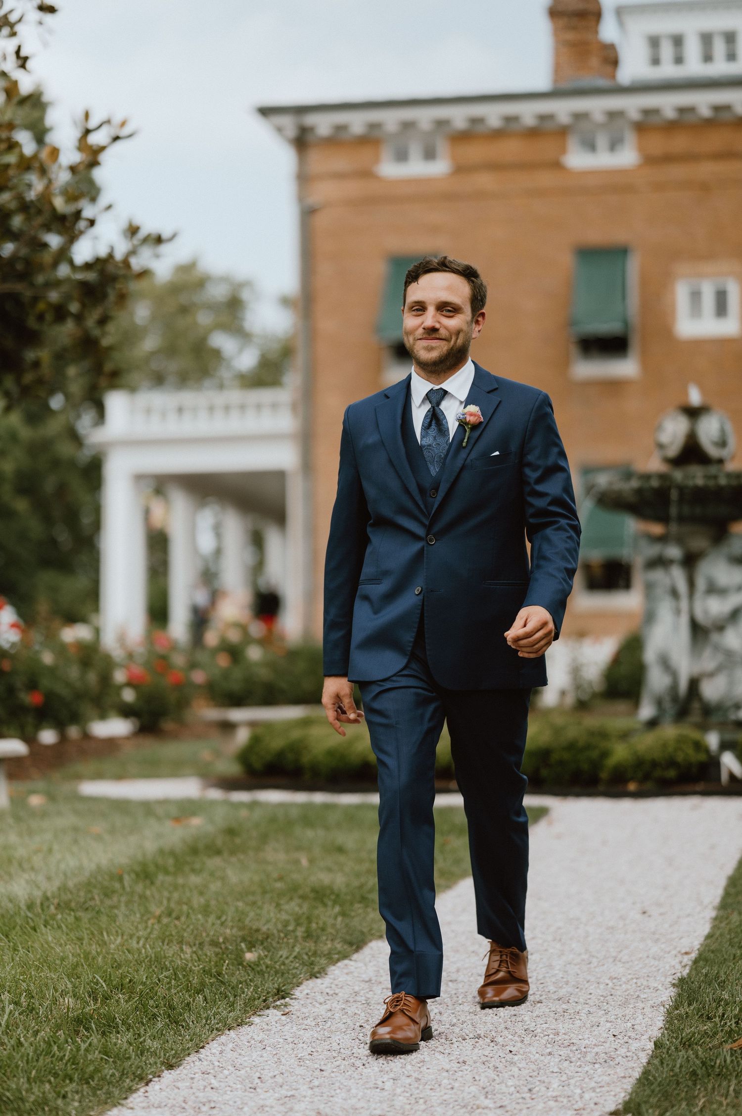 A groom in a navy blue suit walks along a garden path at an elegant venue.