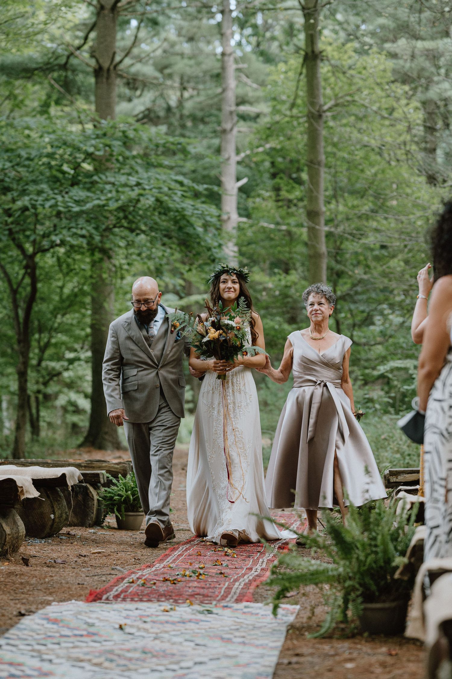 Wedding party walks down a rustic aisle decorated with a red carpet in a forest setting.