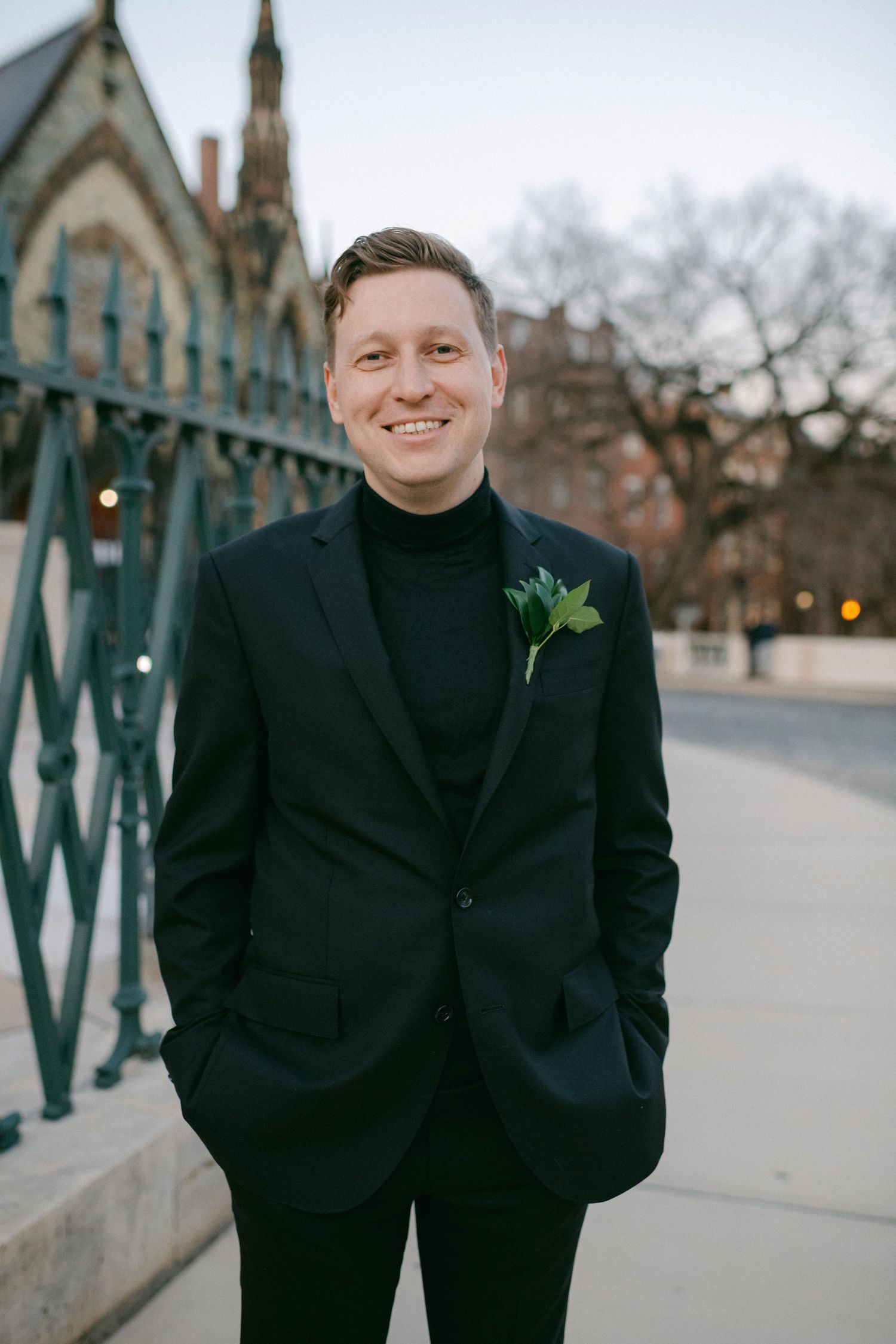 Wedding portrait of a groom in a black suit and turtleneck with boutonniere standing outside historic architecture.