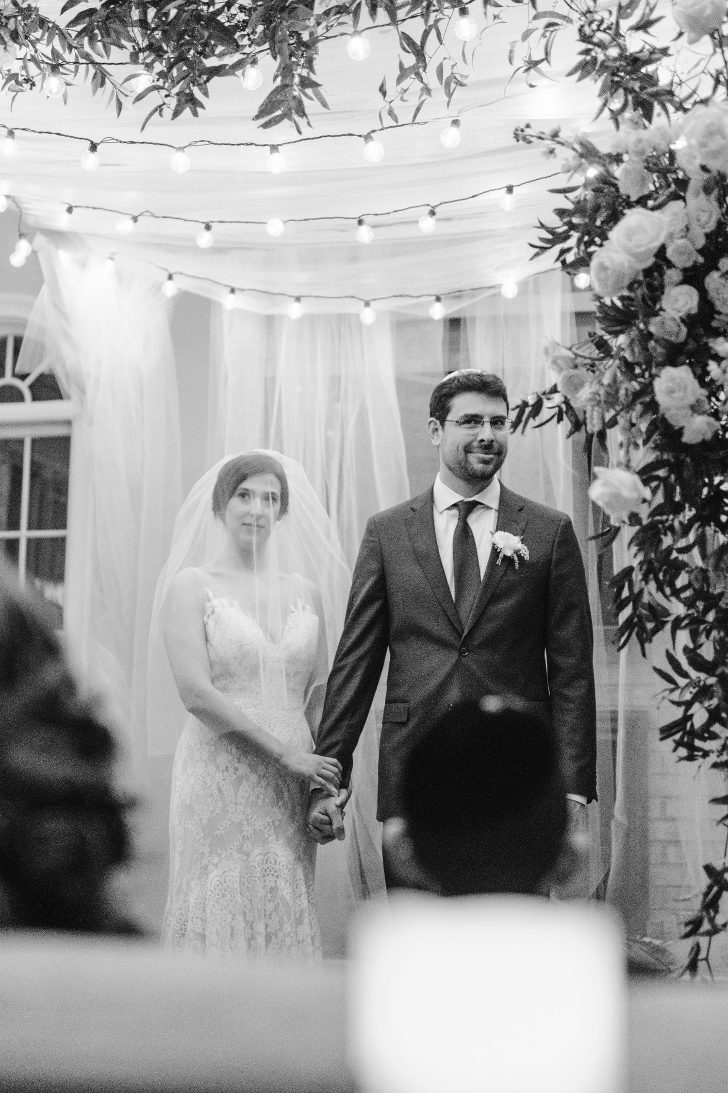 Black and white photo of wedding ceremony under a floral arch with twinkling lights and white drapes.