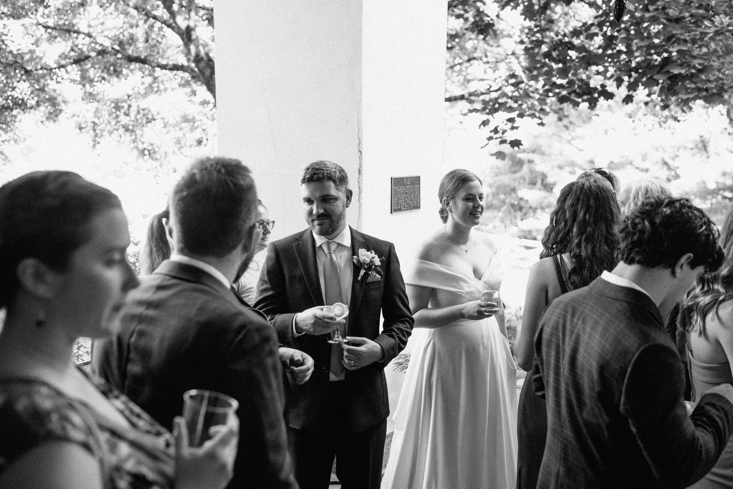Wedding reception guests mingle outside under trees during daytime celebration.