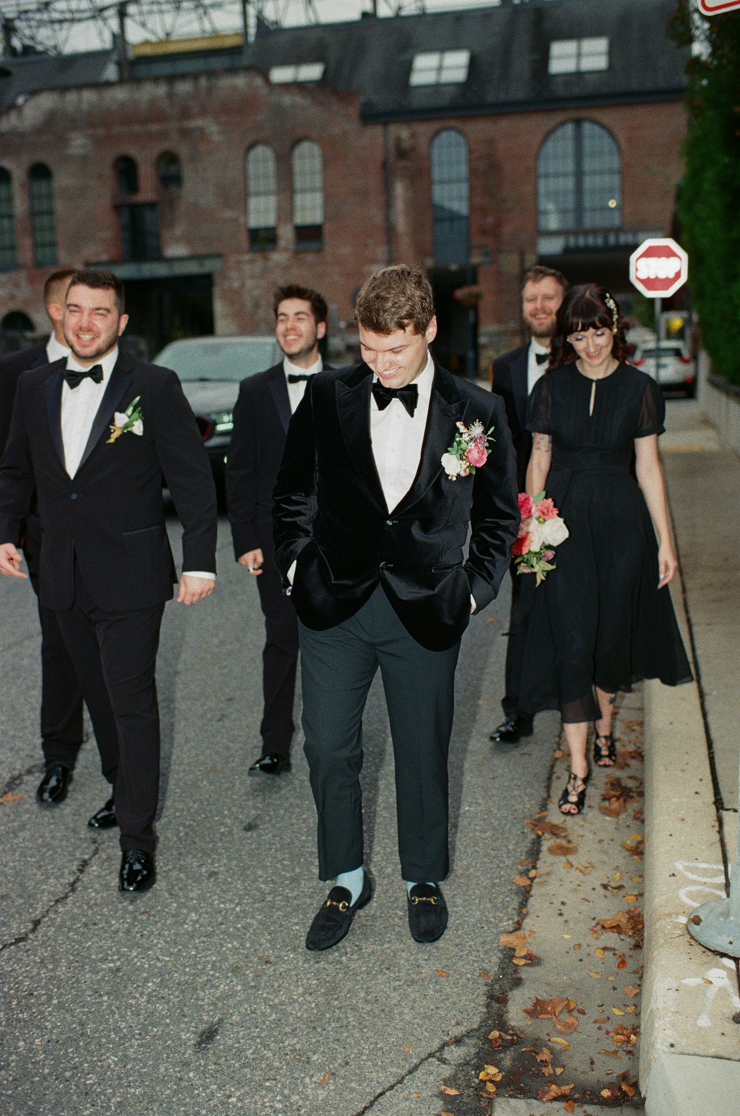 Wedding party in formal attire walks along an urban sidewalk near a brick building.