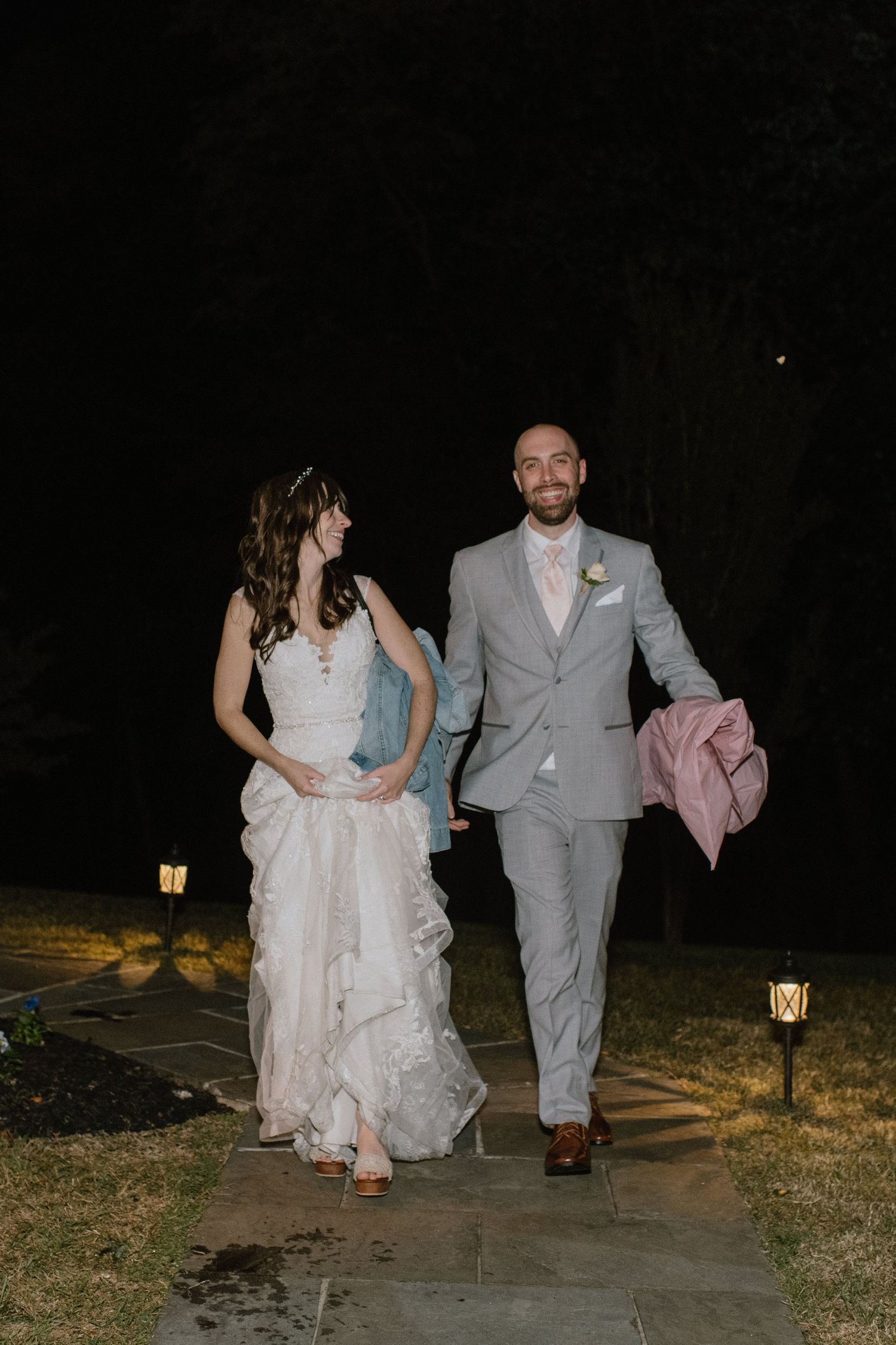 Couple walking along a lit garden path at night after their wedding celebration.