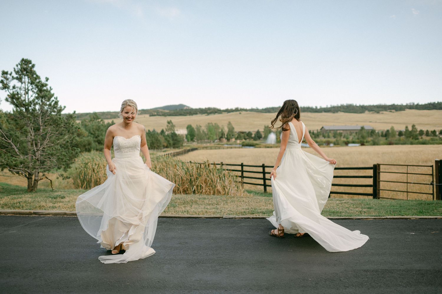 Two people in wedding dresses twirl on a road with rolling hills in the background.