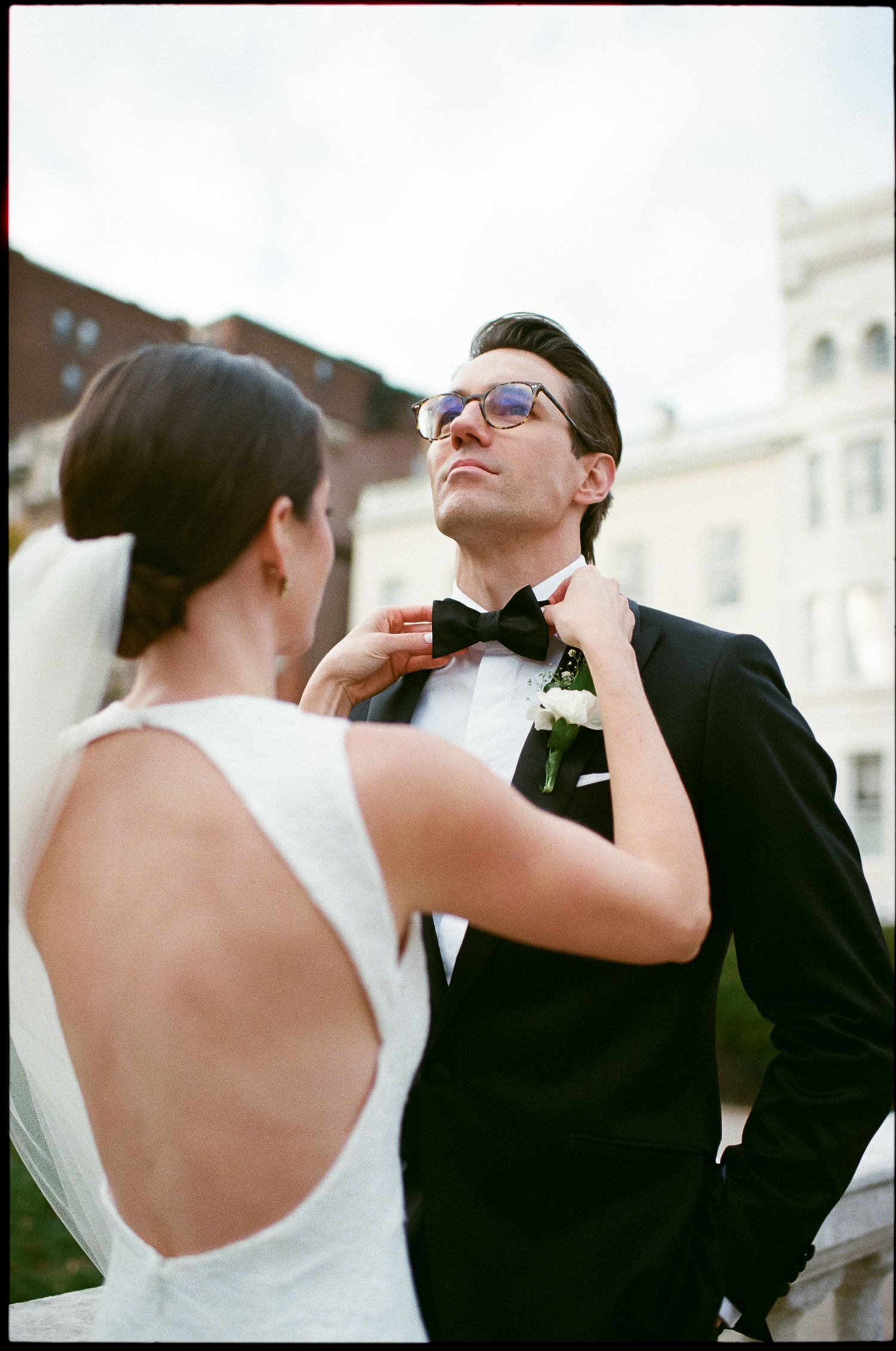 Stylish couple shares moment as bride adjusts groom's bowtie against urban backdrop.