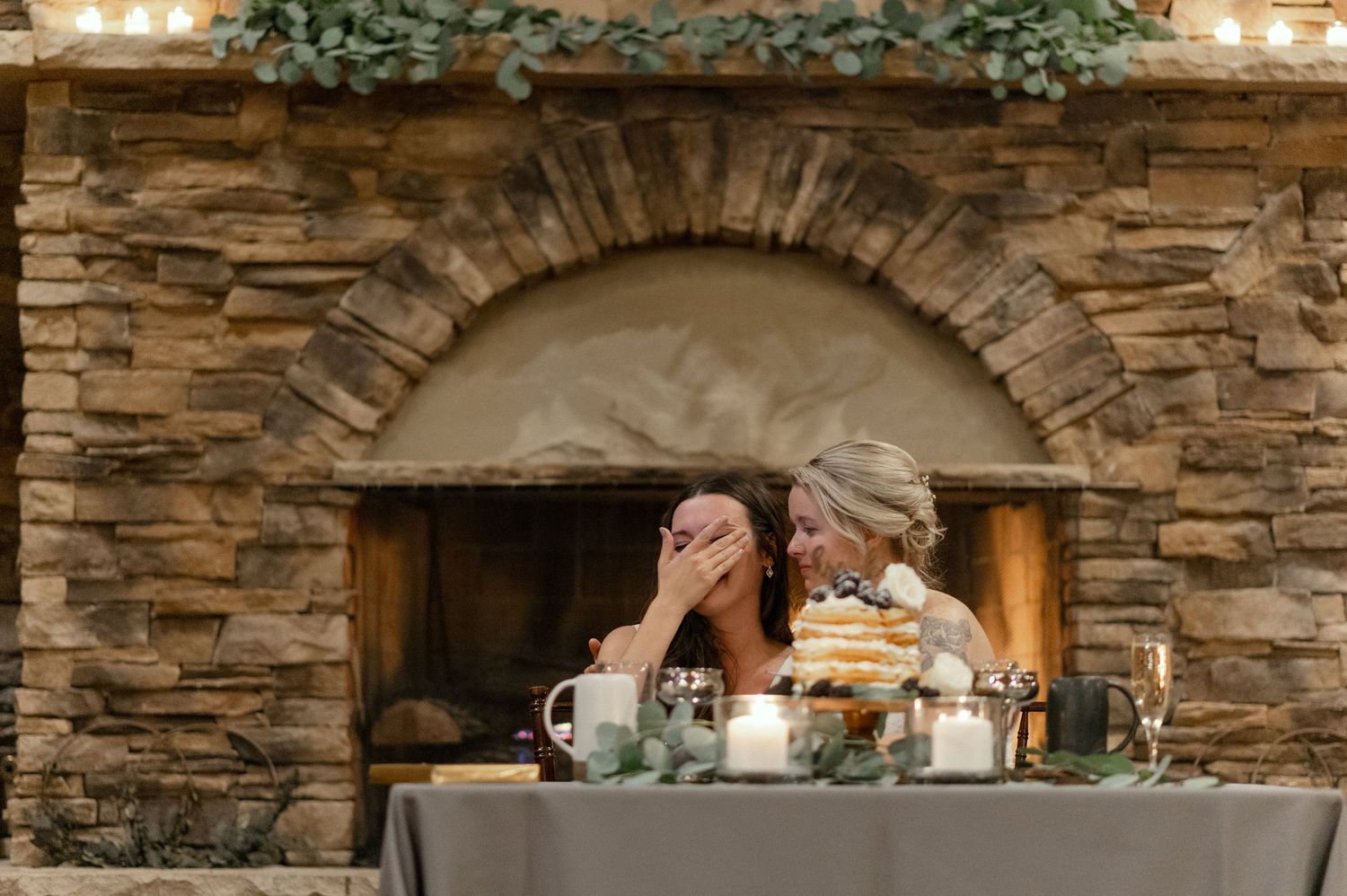 Wedding reception table setup in front of a stone fireplace with decorative greenery.