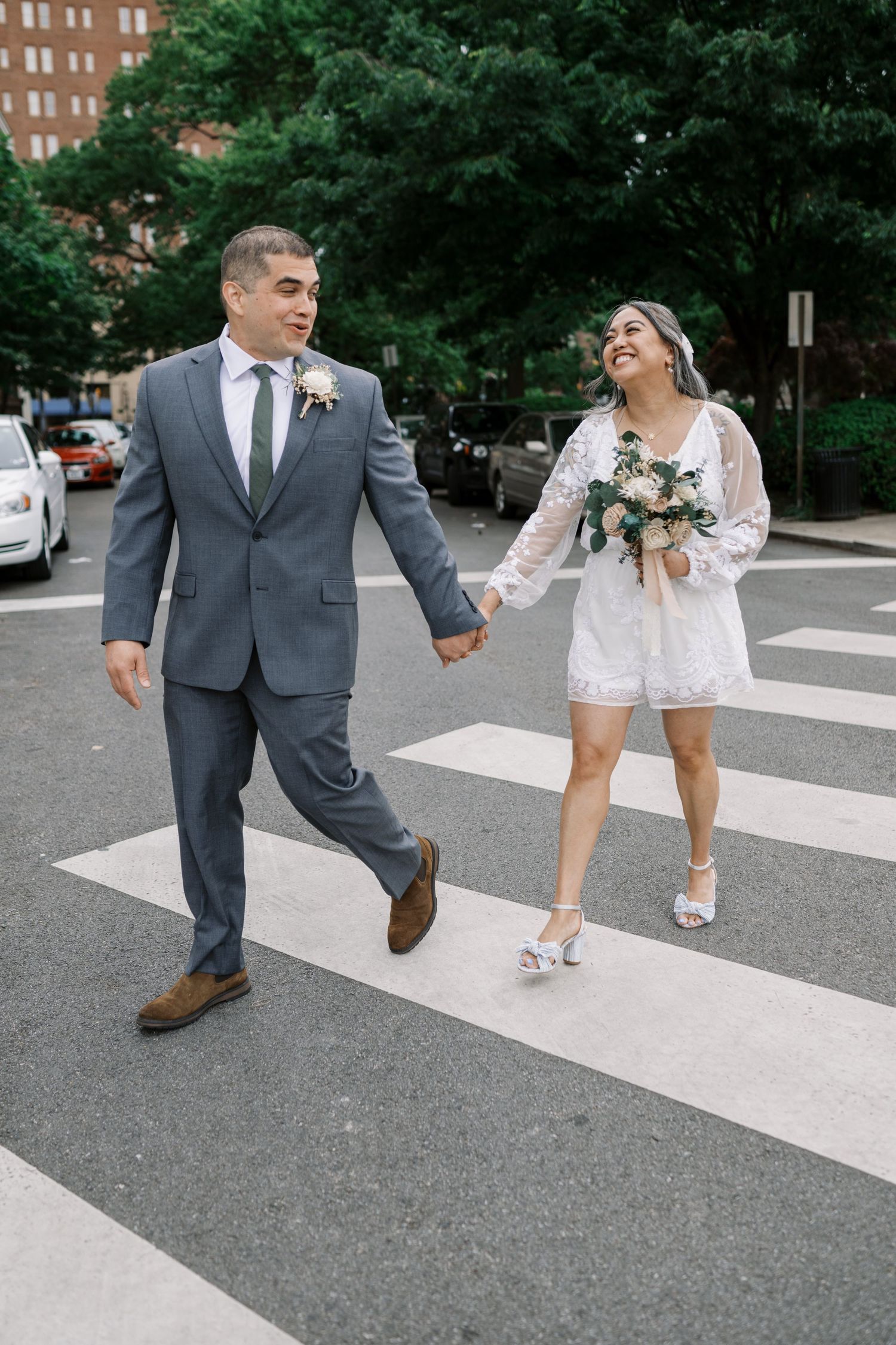 A couple crosses a city street together while holding hands and carrying flowers.