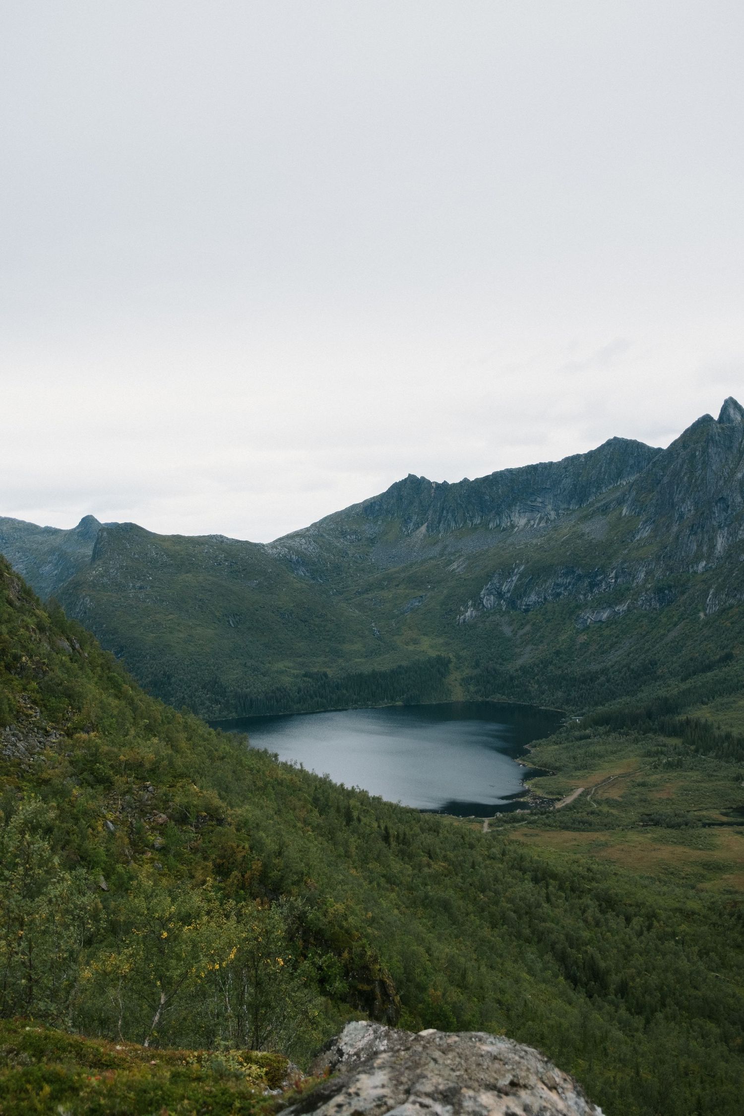 Aerial view of a serene mountain lake nestled among green hills and rugged peaks under an overcast sky.