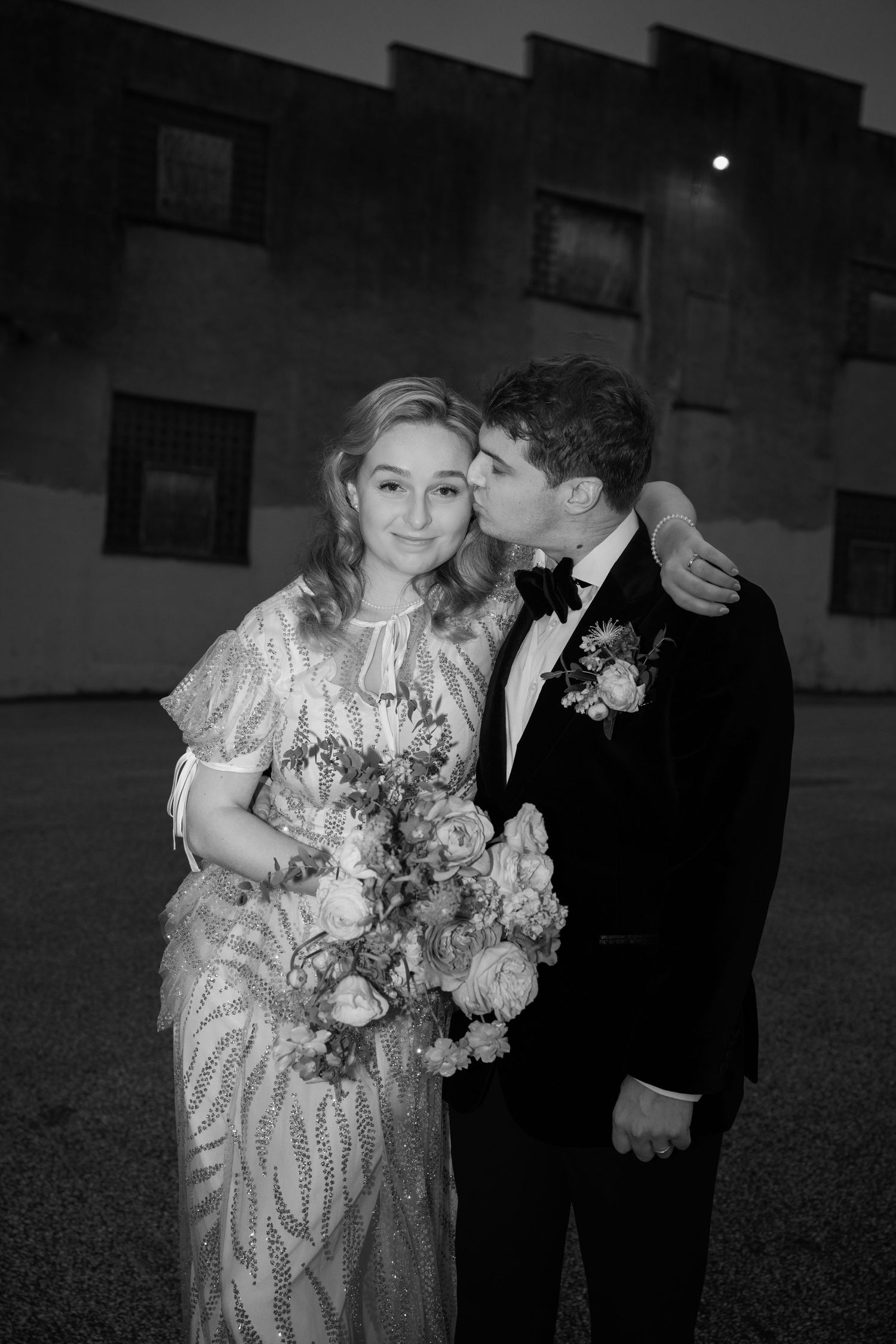 A romantic black and white portrait of a couple with bouquet against urban backdrop.