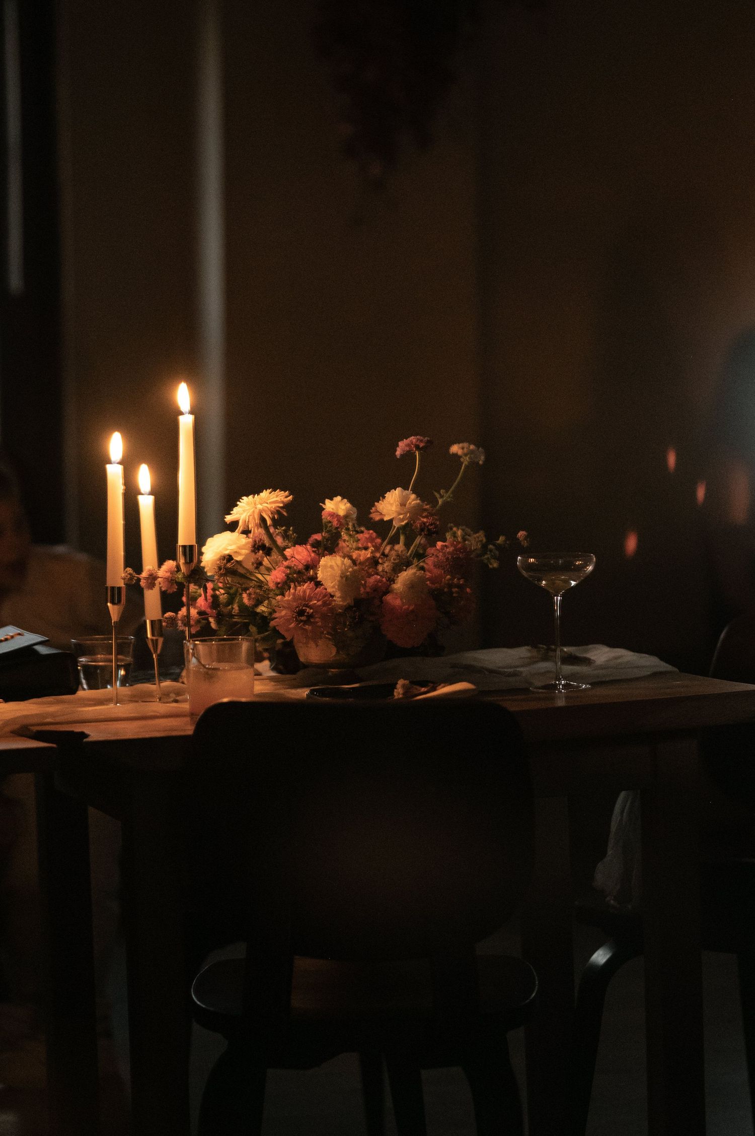 Romantic candlelit dinner table with floral centerpiece, glowing taper candles, and wine glasses in a dark, moody setting.