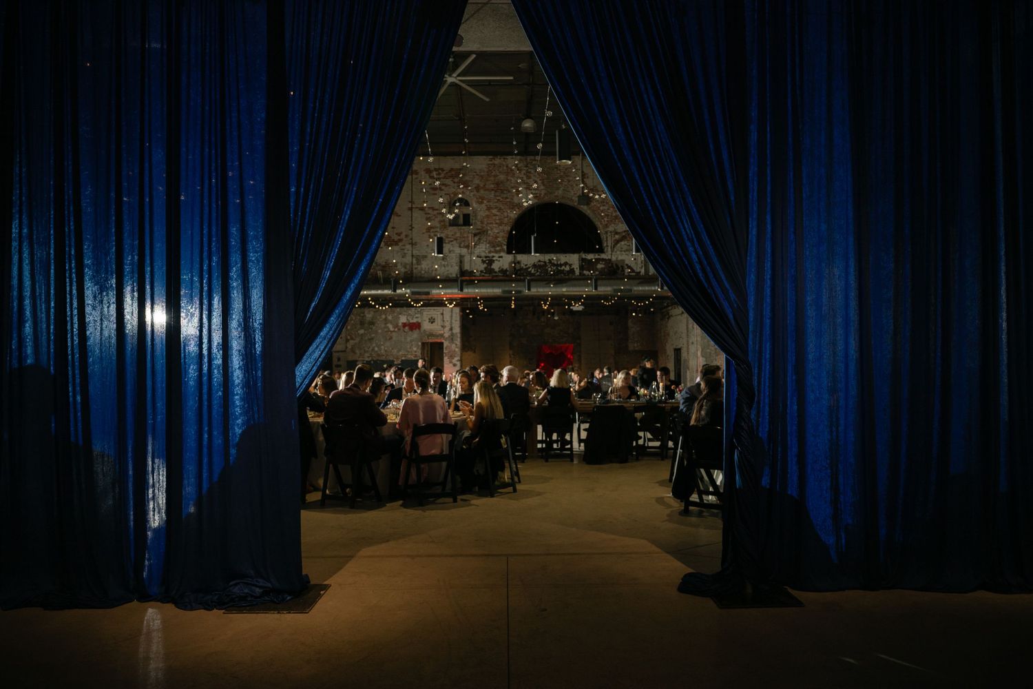 Elegant event venue with blue velvet curtains framing a dimly lit reception hall with guests seated at round tables.