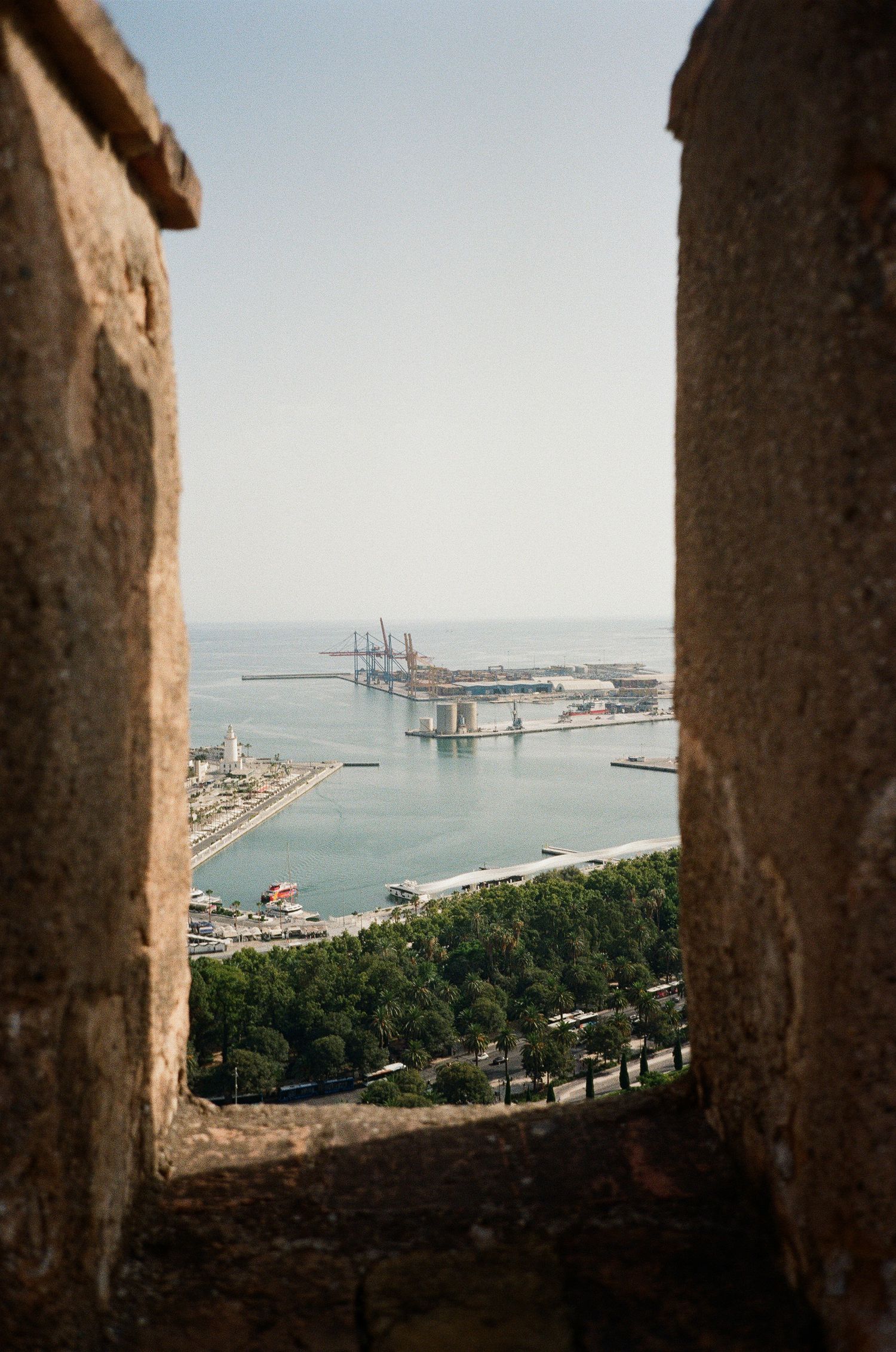 View of a harbor through stone archway frames with ships and coastal buildings visible in distance.