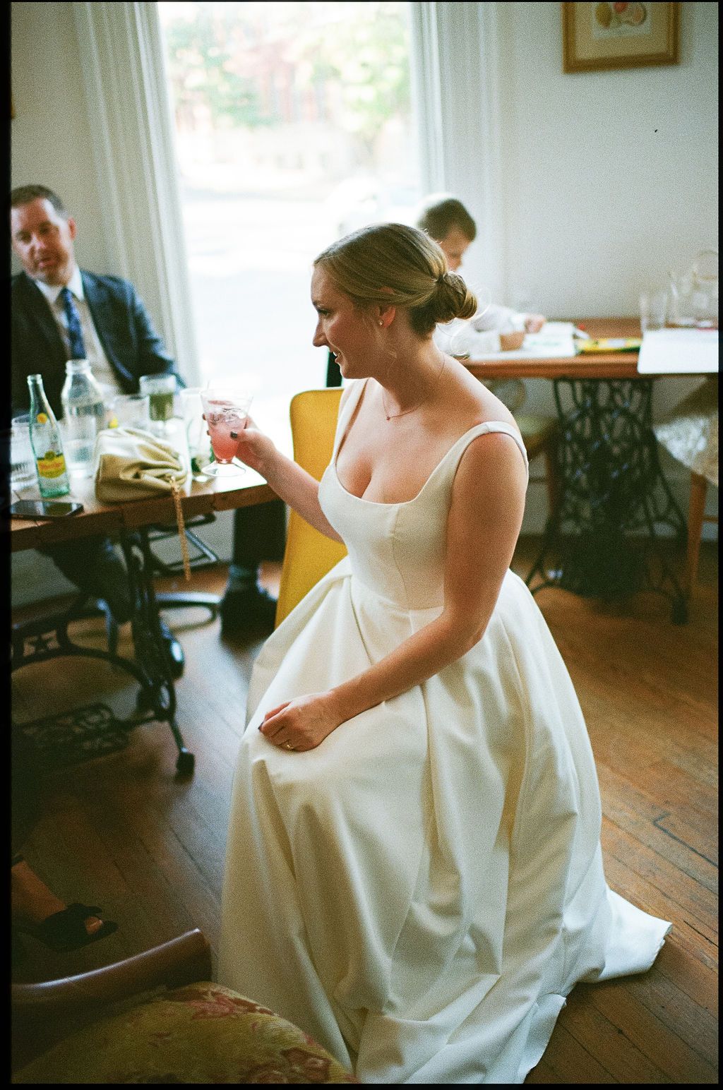 A bride in a classic white dress laughs while sitting in a vintage-style room with wooden floors.