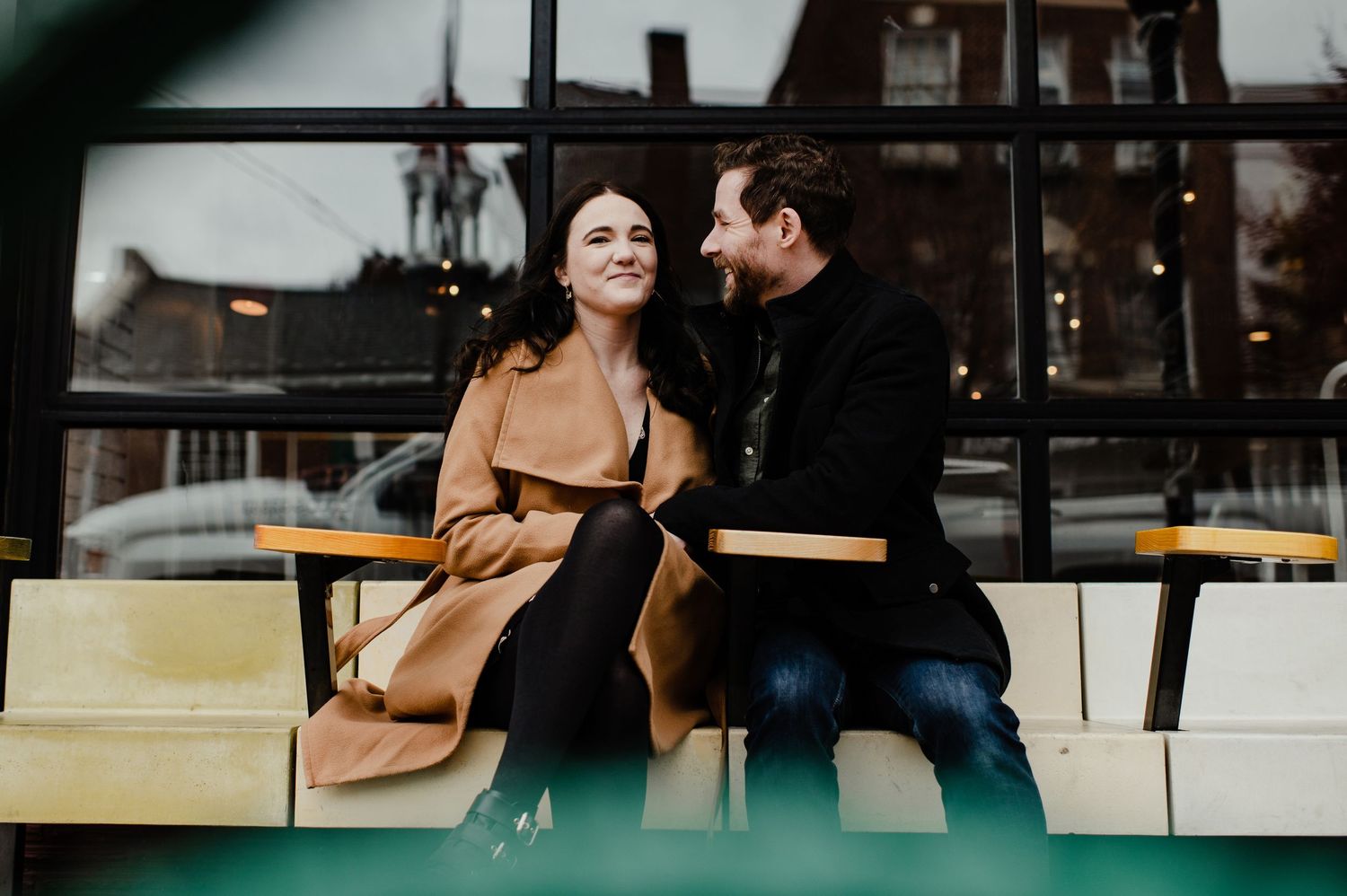 Couple sitting together on a bench outside a cafe, smiling and sharing an intimate moment in winter coats.