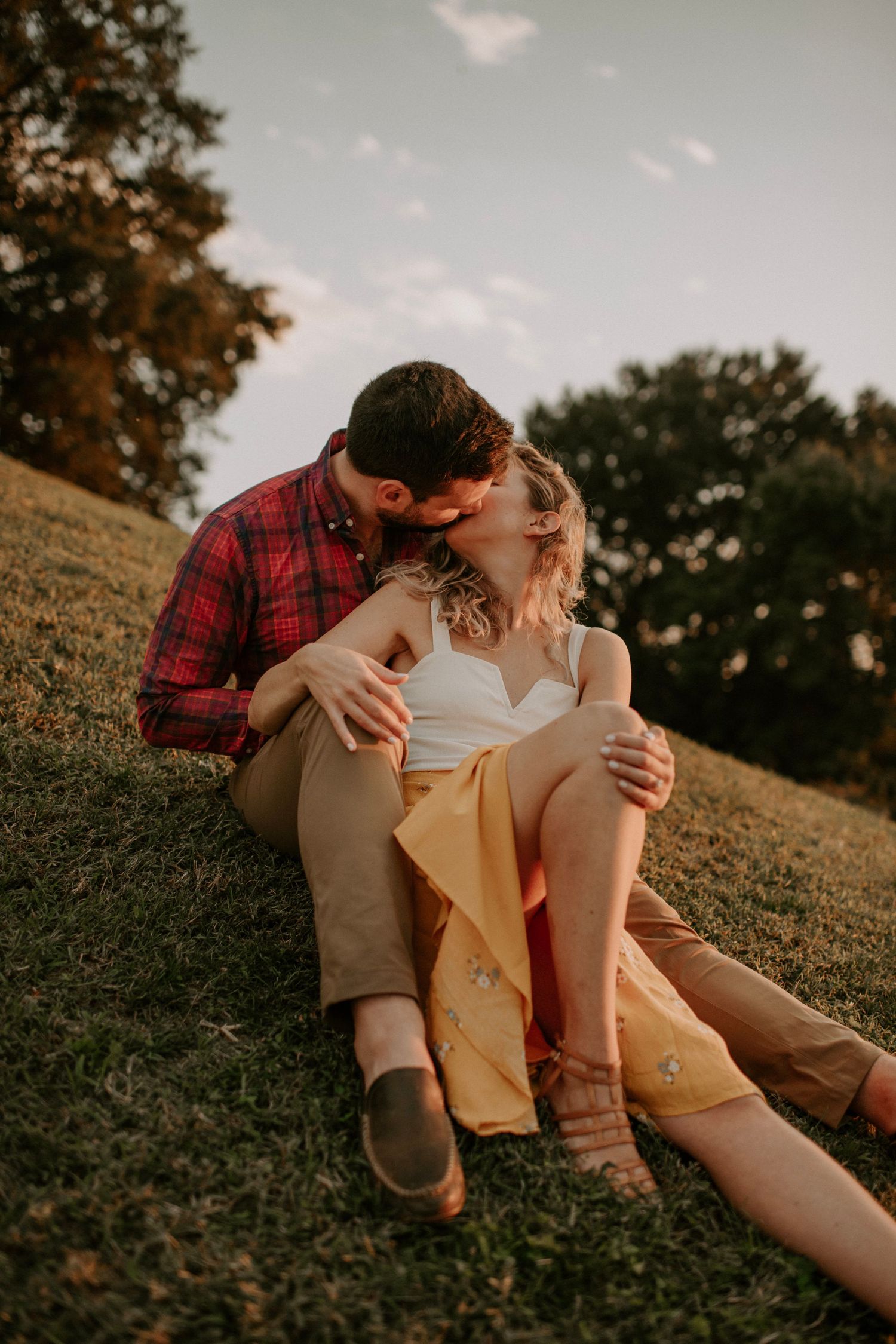 A couple shares a romantic kiss while sitting on a grassy hill, the woman in a yellow skirt and white top.
