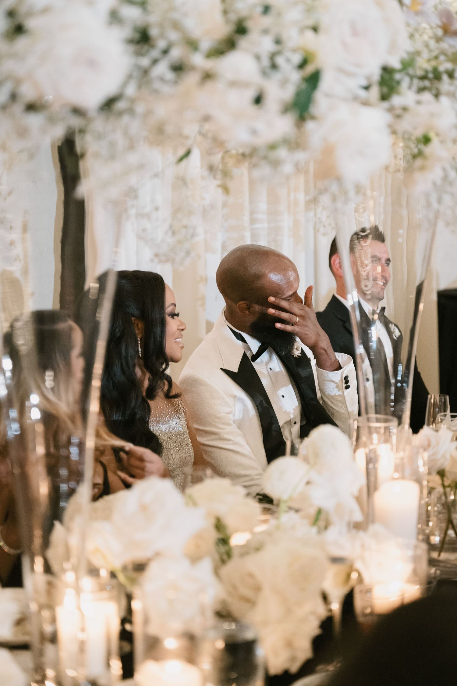 Emotional couple seated at elegant wedding reception table adorned with white floral arrangements and candlelight.