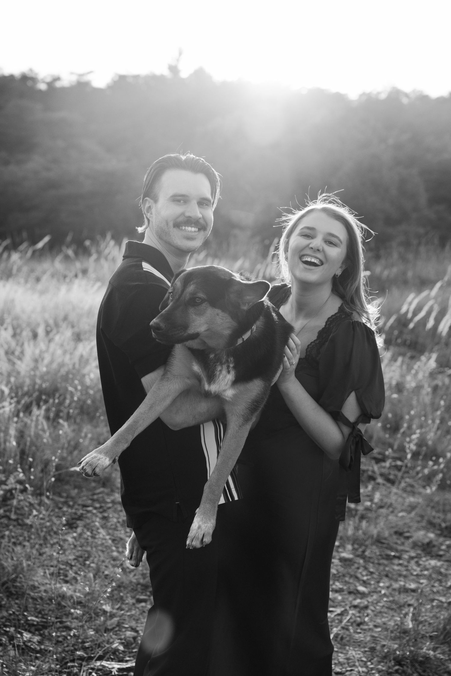 A couple shares a joyful moment with their black dog in a sunlit field at golden hour.