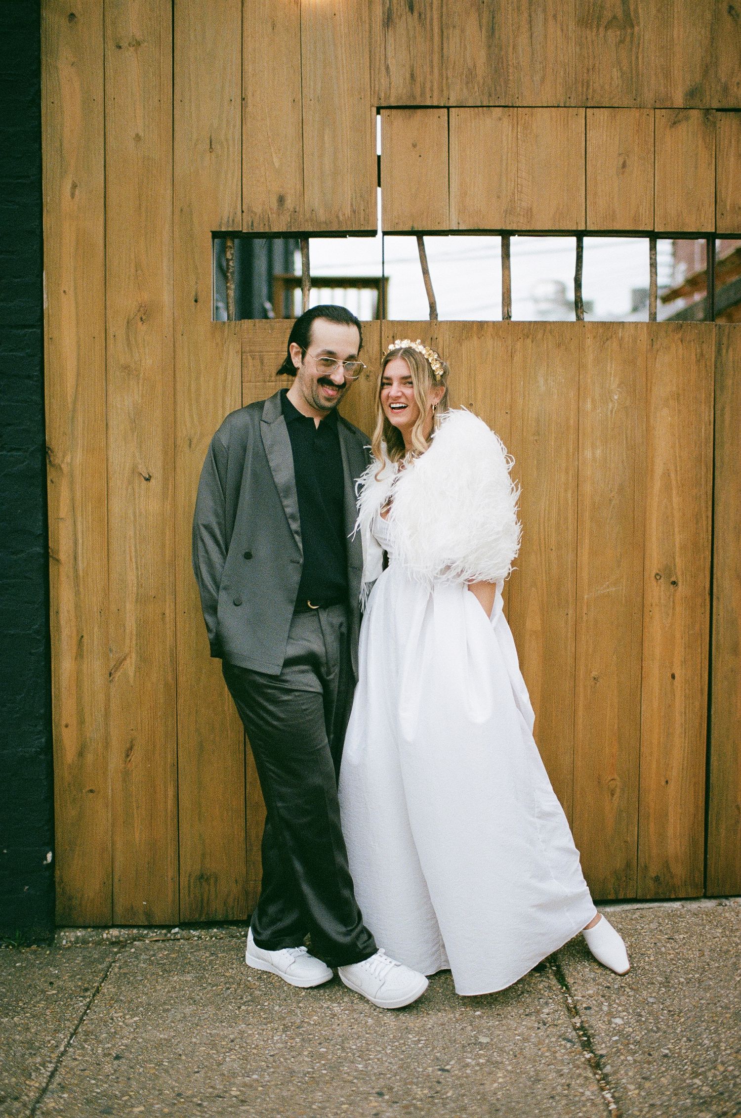 A casual wedding couple posing against a rustic wooden wall, with the bride wearing a white fur wrap.