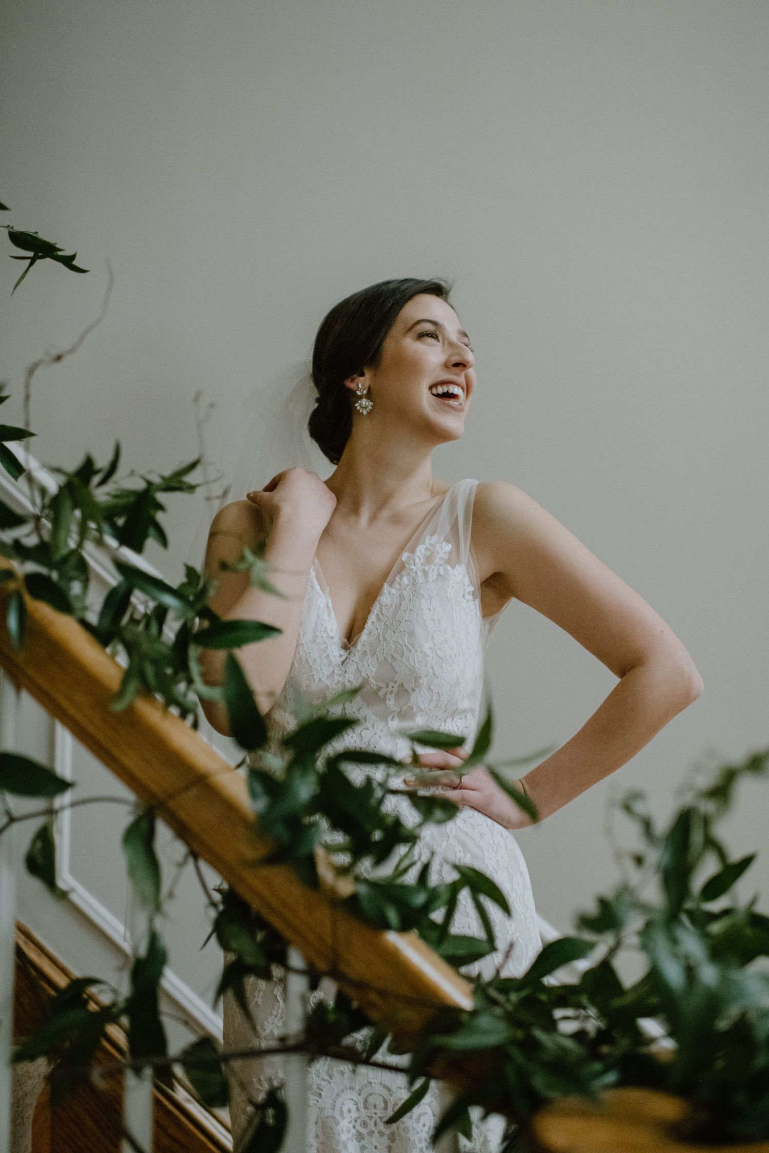A bride in a lace wedding dress laughs joyfully among decorative greenery on a staircase.