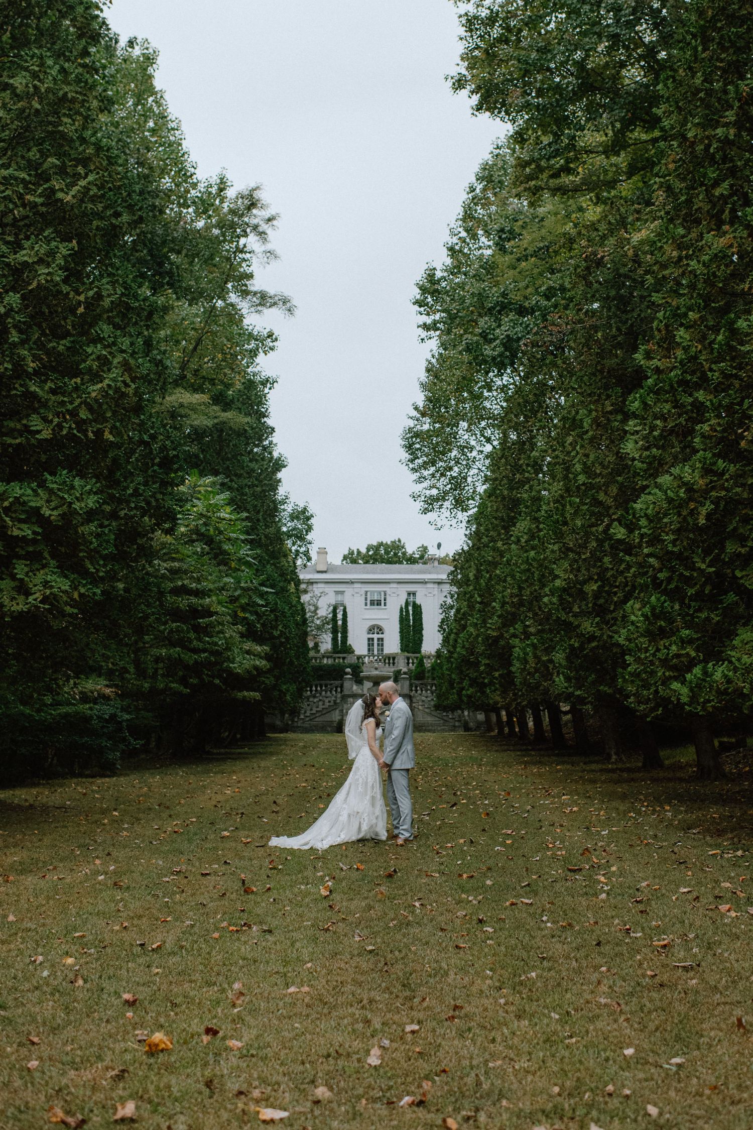 Wide shot of newlyweds standing in front of a classical mansion at the end of a tree-lined path.