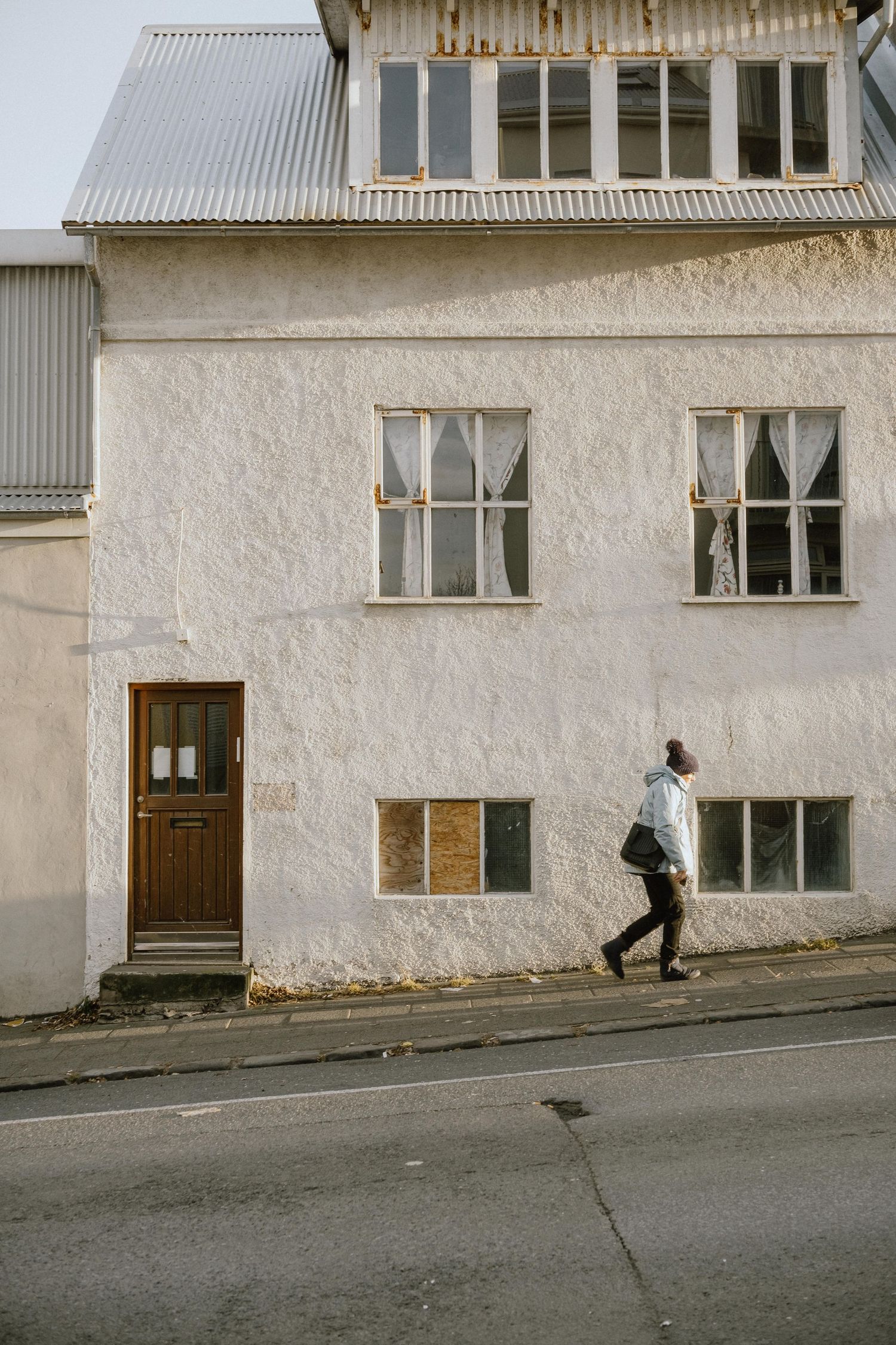 Person walking past a white stucco building with vintage windows on a sloping street.