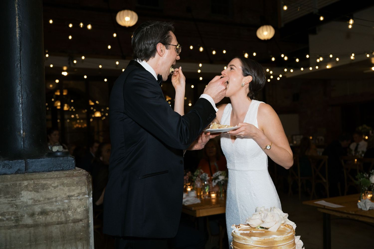 Newlyweds share a playful moment while feeding each other wedding cake under romantic string lights.