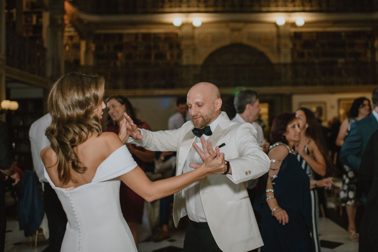 A couple in white formal attire dances together at an elegant indoor reception venue with guests in background.