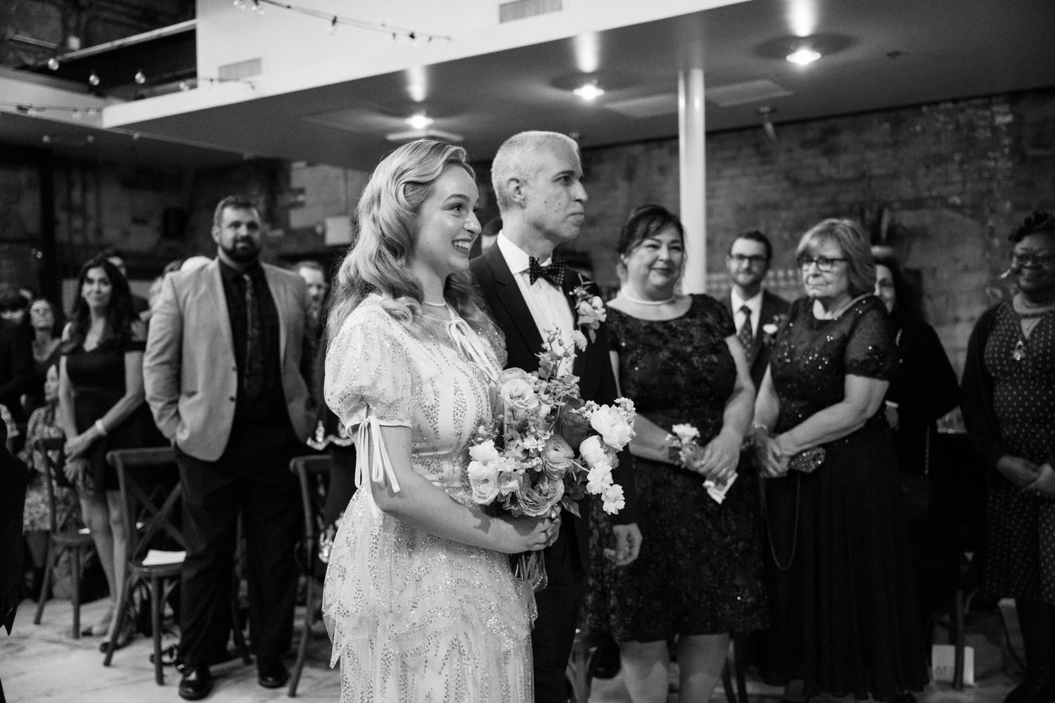 Black and white photo of couple during intimate indoor wedding ceremony with guests looking on.
