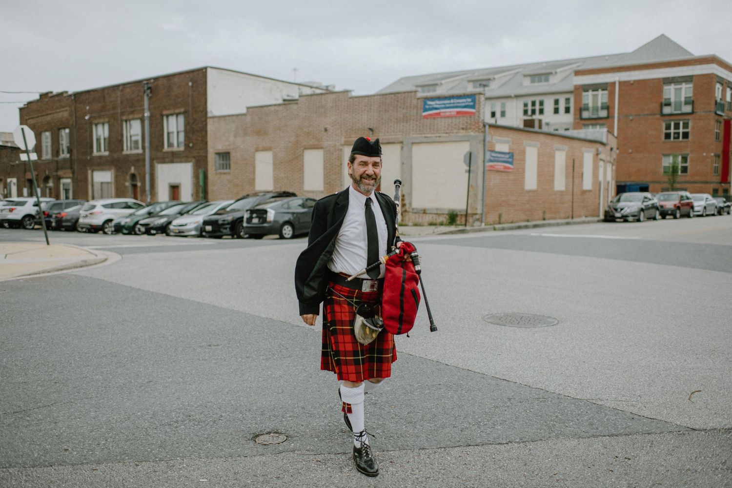 Person wearing traditional Scottish kilt walking across parking lot carrying bags.