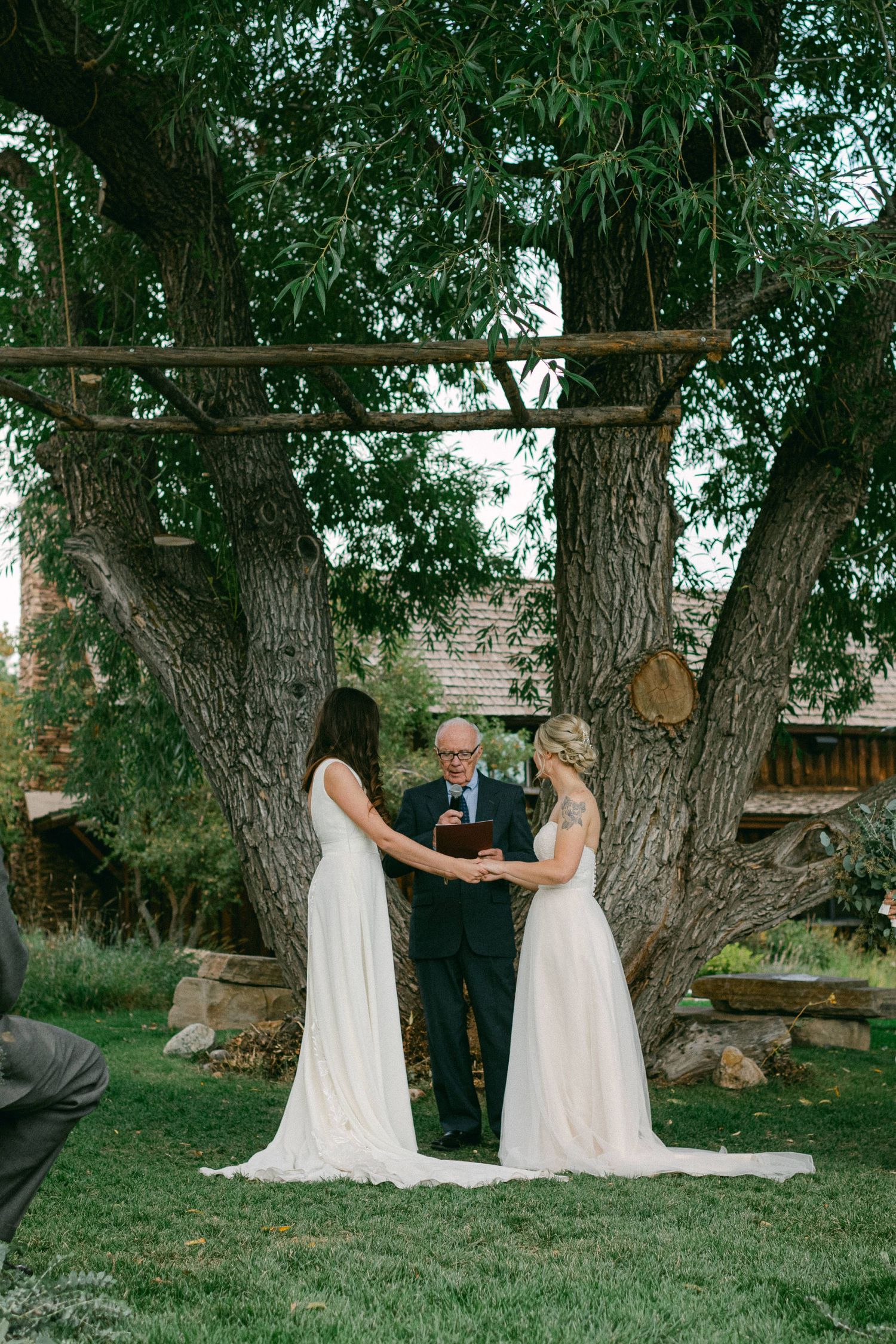 Wedding ceremony under a rustic wooden arch between large trees with flowing white dresses in view.
