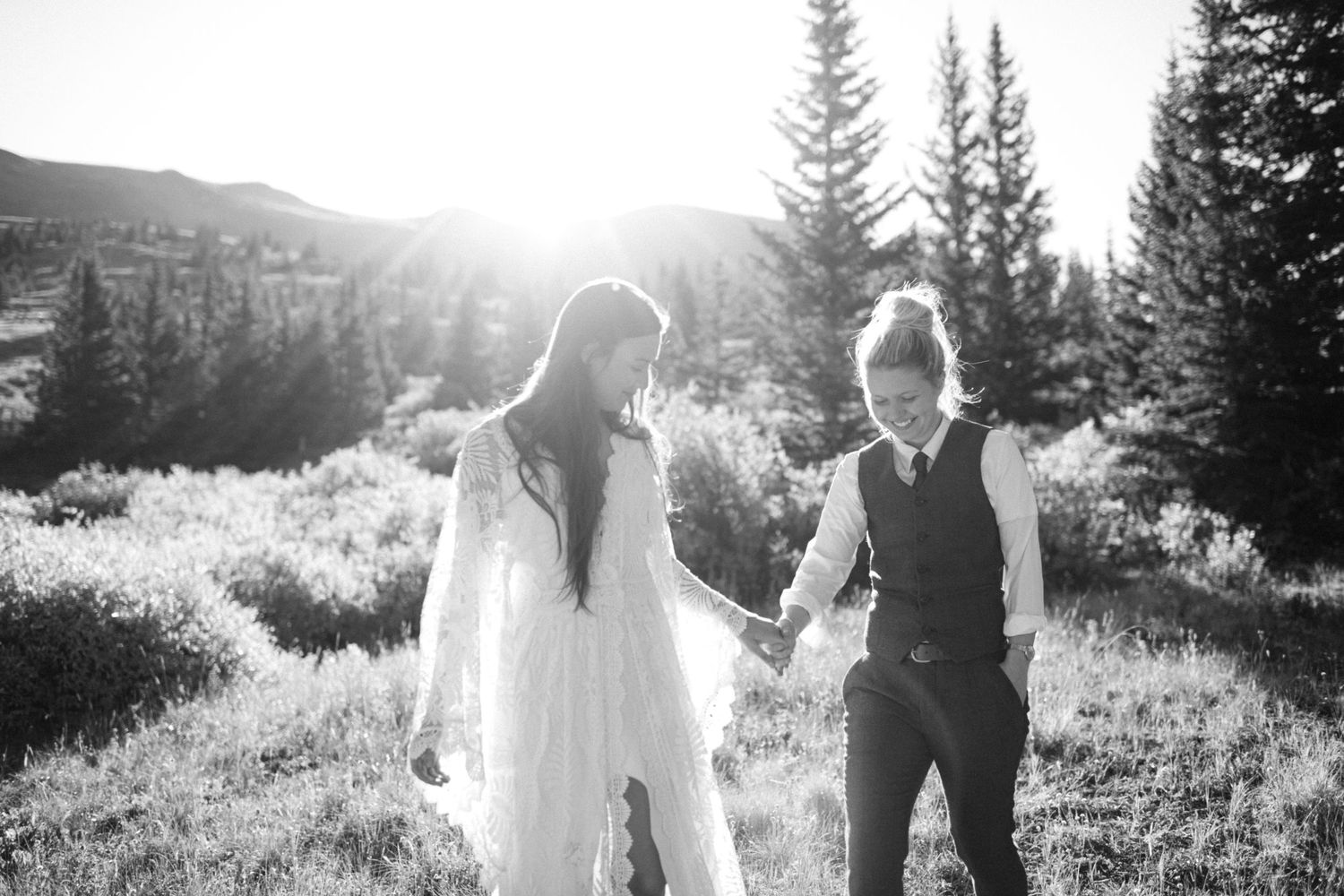 A black and white photo of two people holding hands in a sunlit mountain meadow.