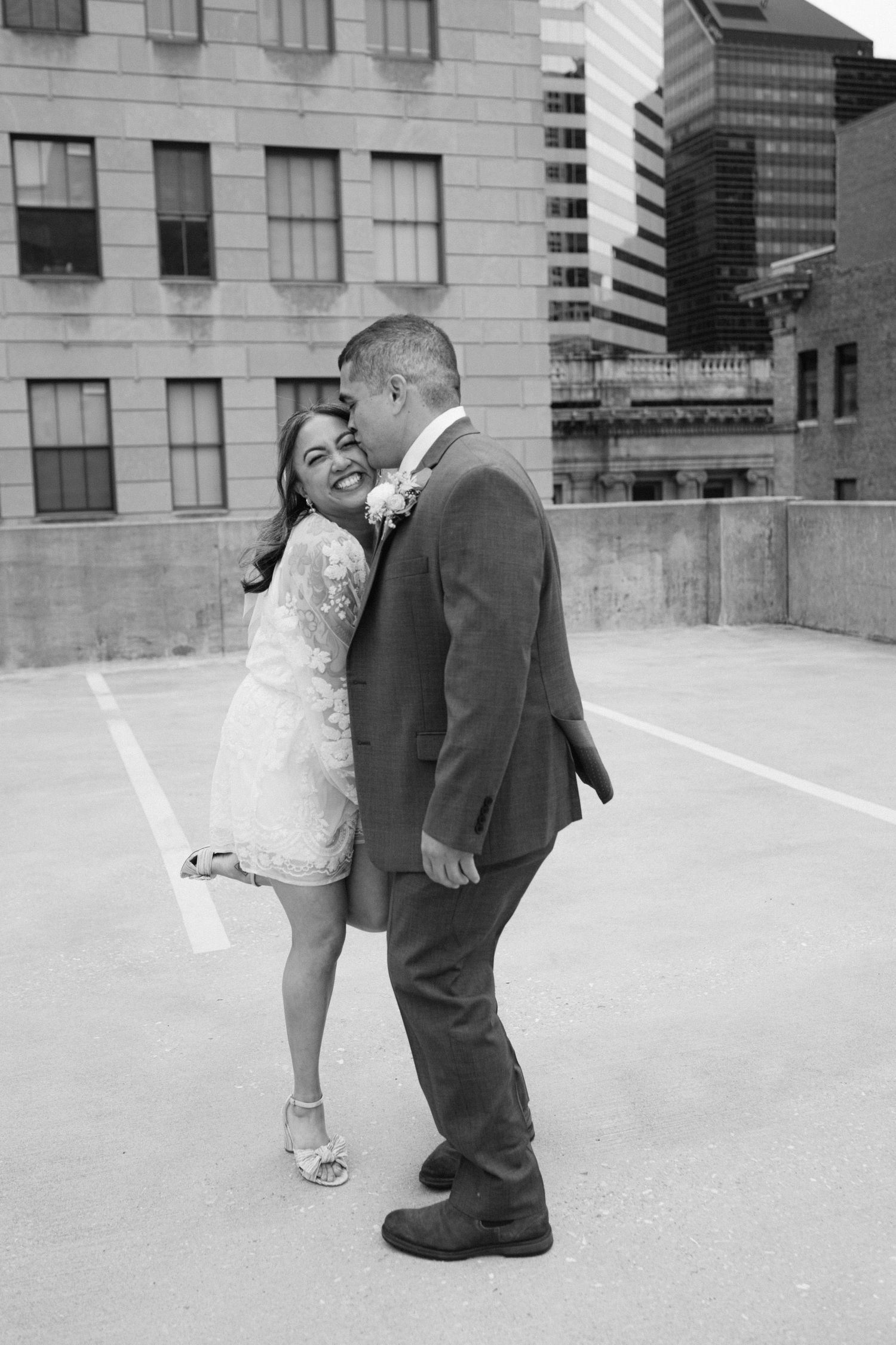 A black and white photo of a couple embracing on a rooftop parking lot in an urban setting.