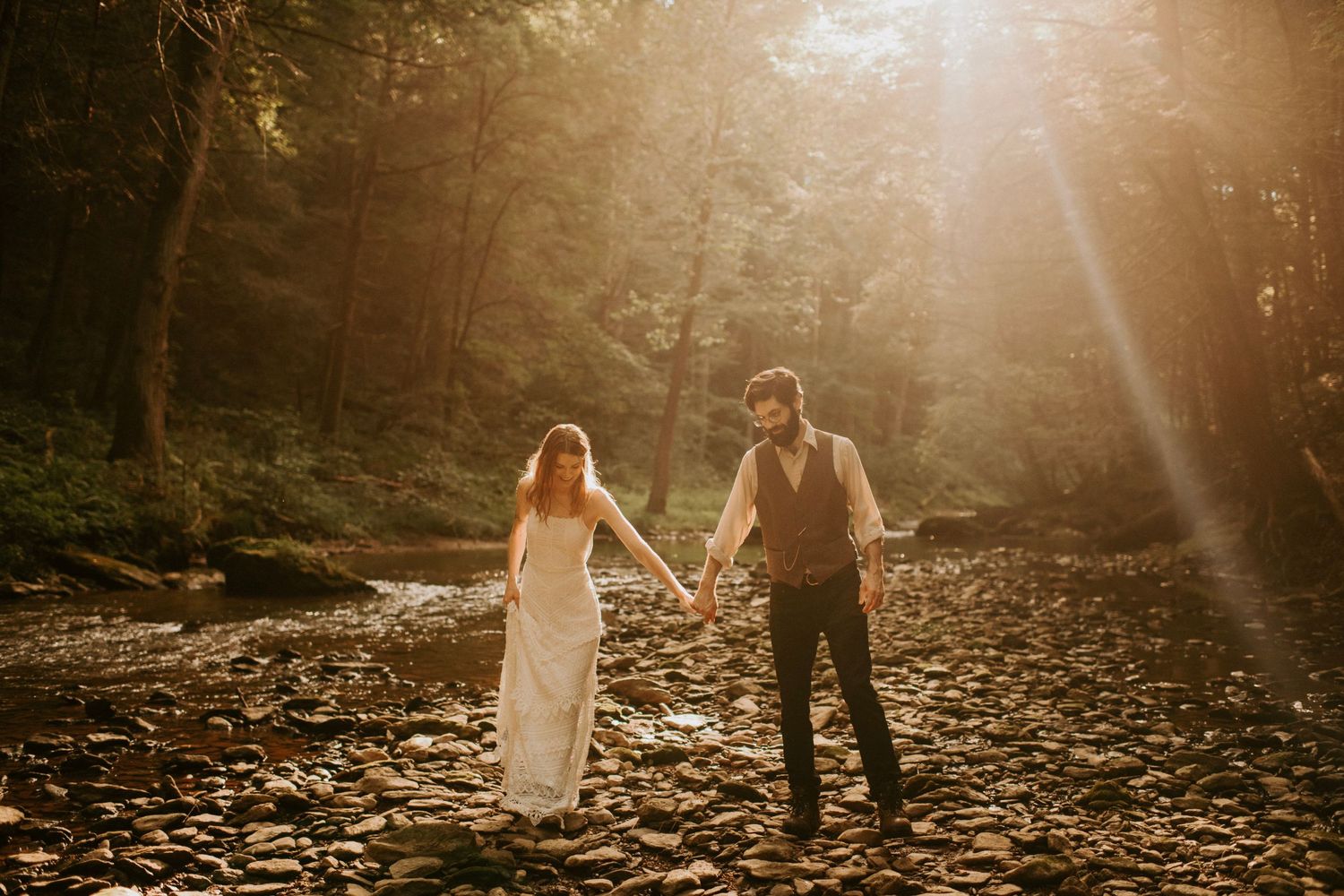 Couple walks hand in hand through golden sunlit forest clearing.