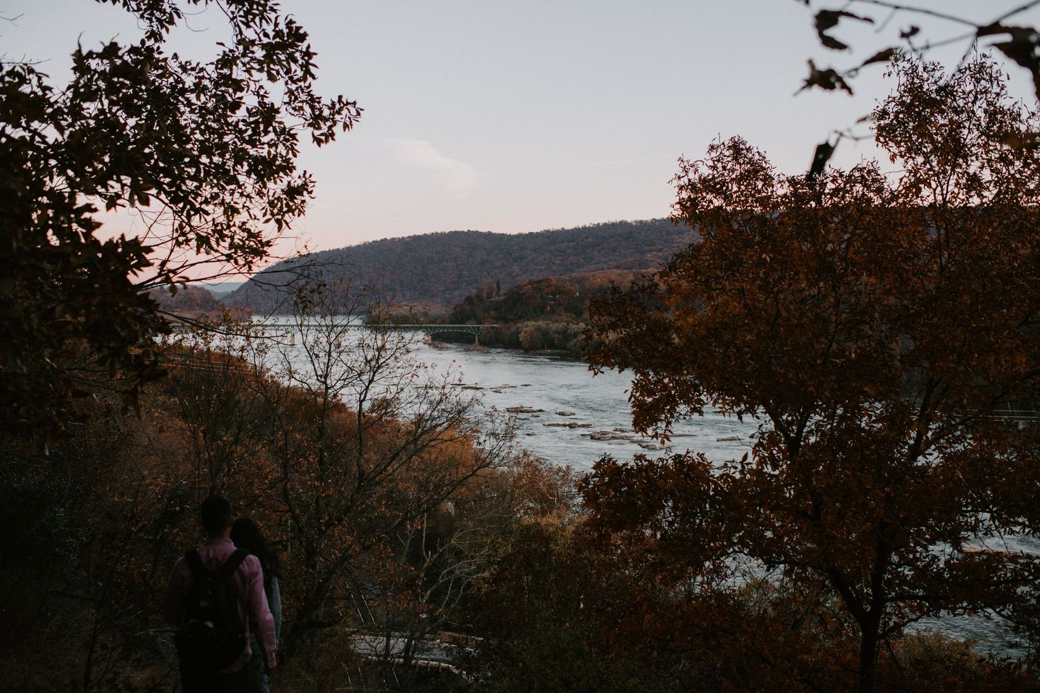 A couple stands silhouetted against a scenic river view at dusk with trees framing the landscape.