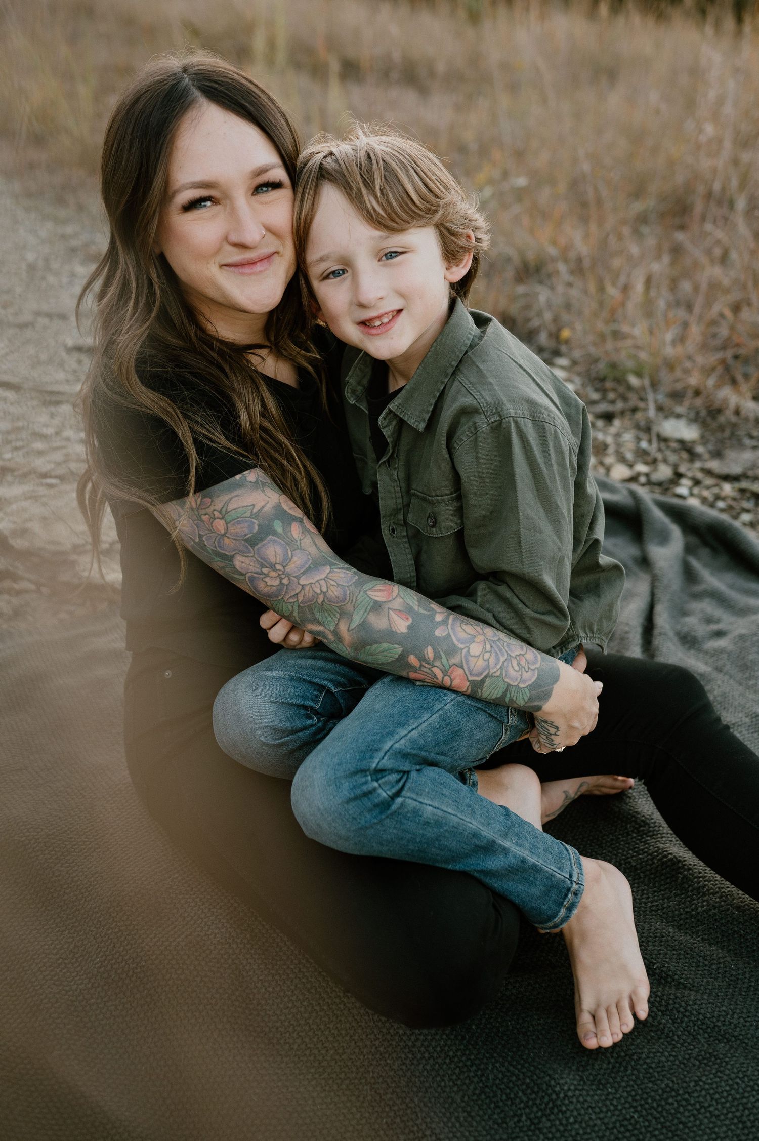 An intimate portrait shows parent and child embracing while sitting on a blanket outdoors at sunset.