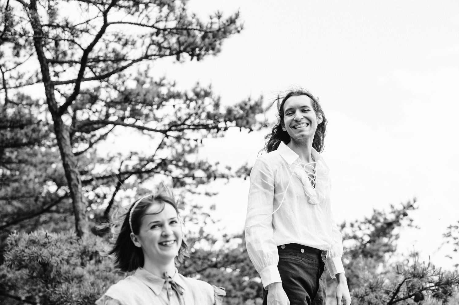 Black and white portrait of two people outdoors with pine trees in background.