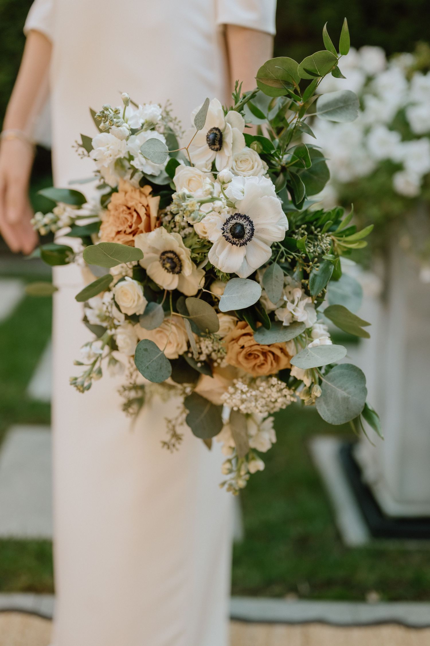 A romantic bridal bouquet featuring white anemones, peach roses, and eucalyptus greens against a white dress.