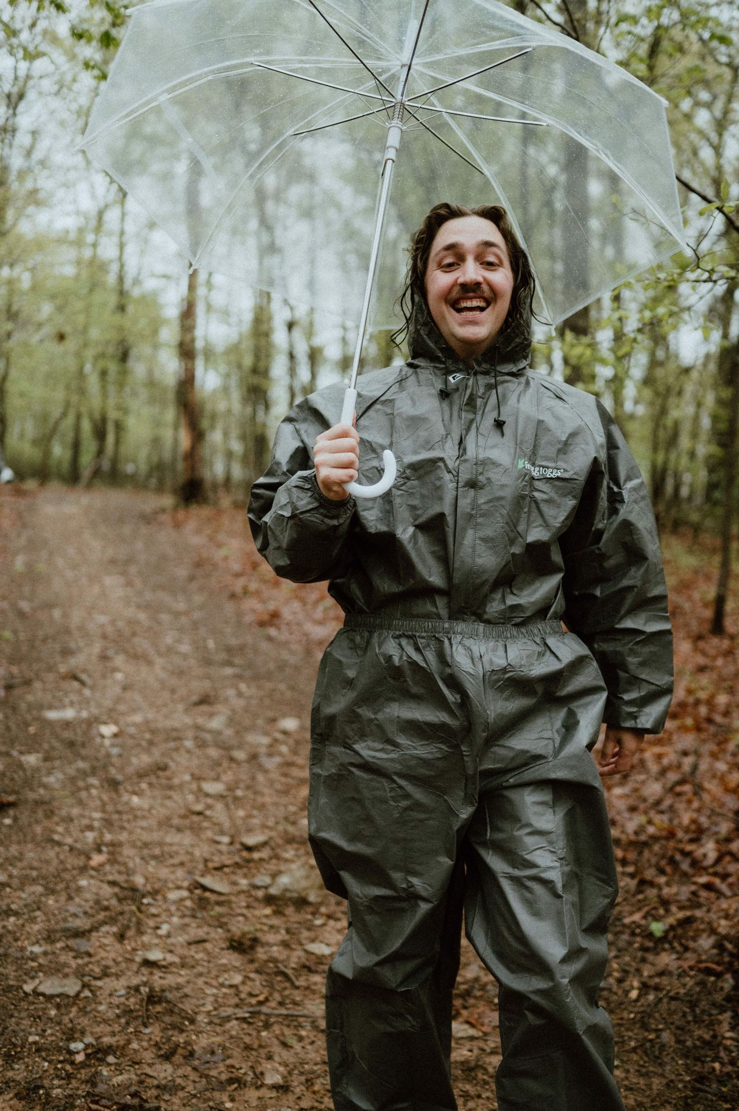 Someone in a full rain suit holds clear umbrella while laughing on a forest trail.