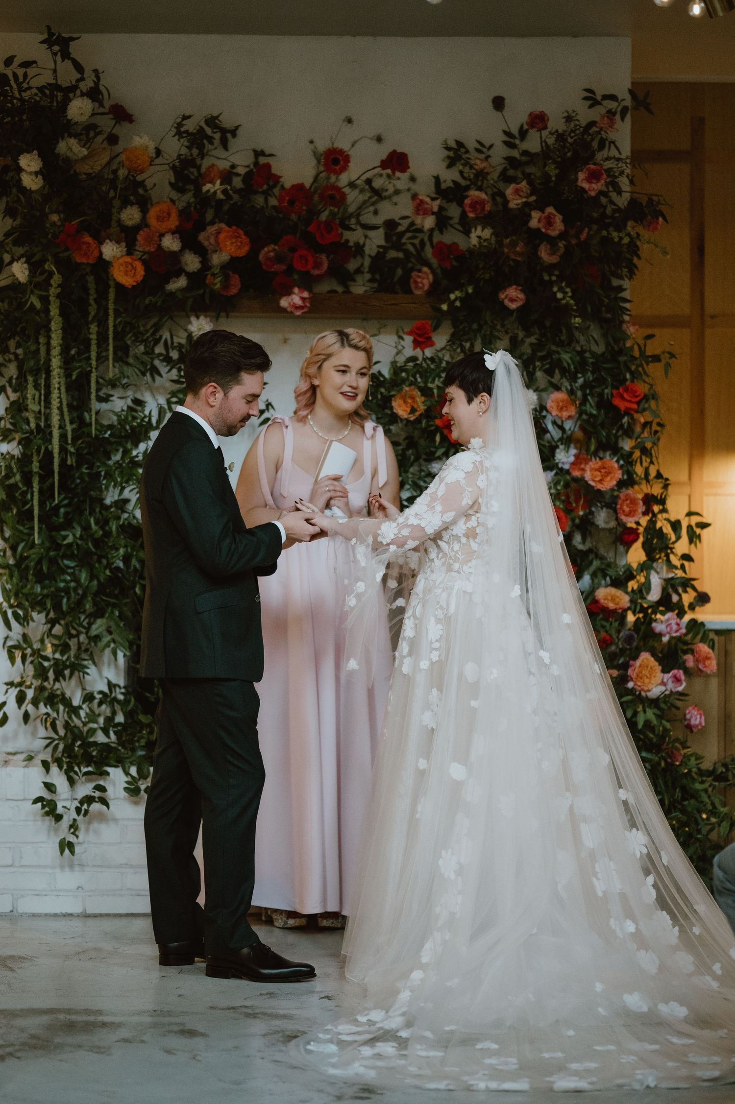 A wedding ceremony takes place under a beautiful floral arch with red and orange blooms.