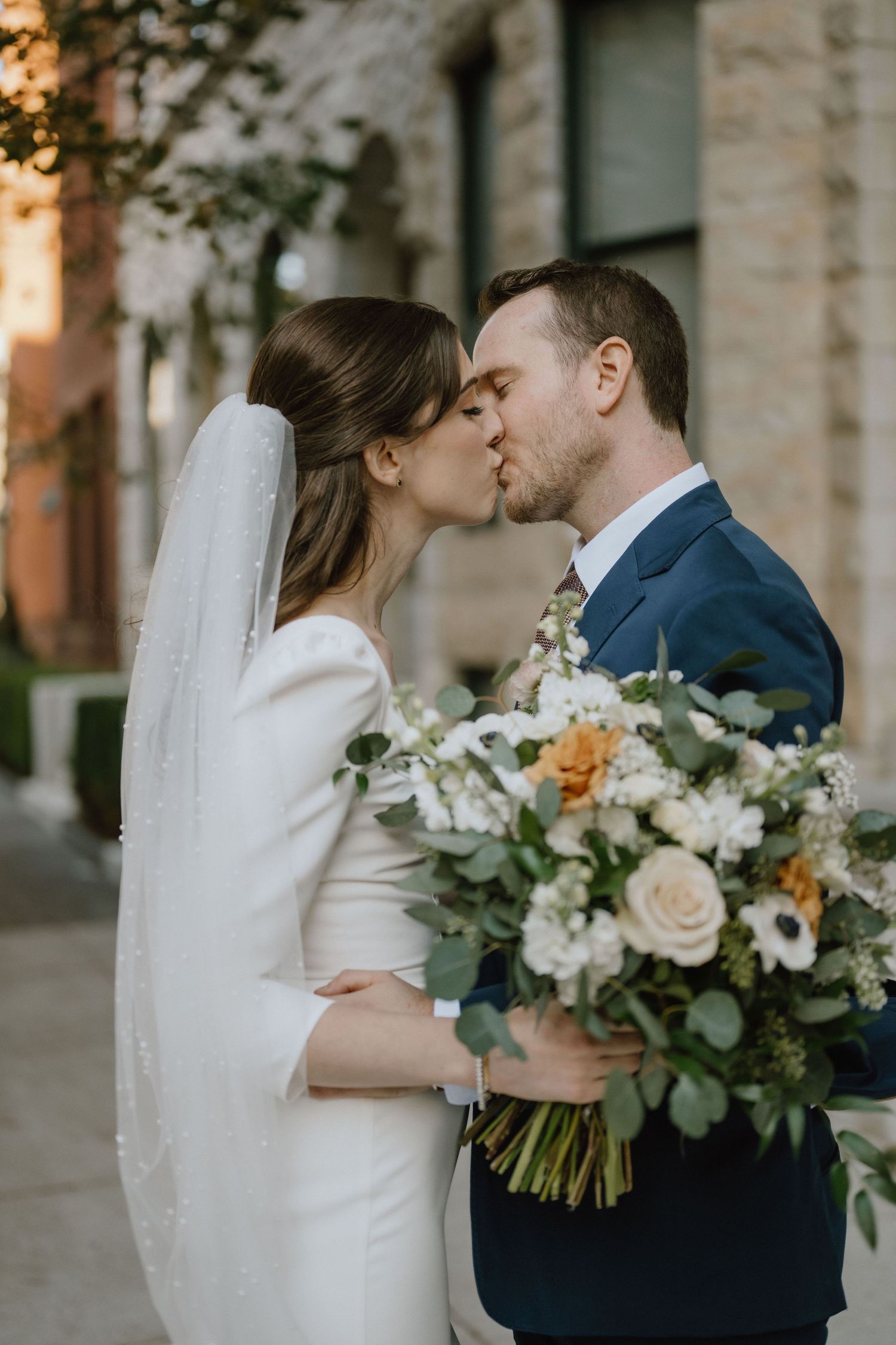 Elegant wedding couple sharing a romantic kiss while holding a lush bouquet of white and peach flowers.
