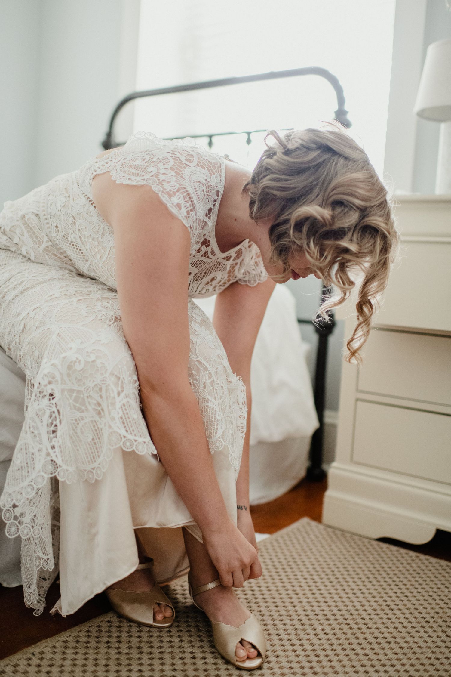 A bride in a lace wedding dress puts on her shoes while sitting on the edge of a bed.