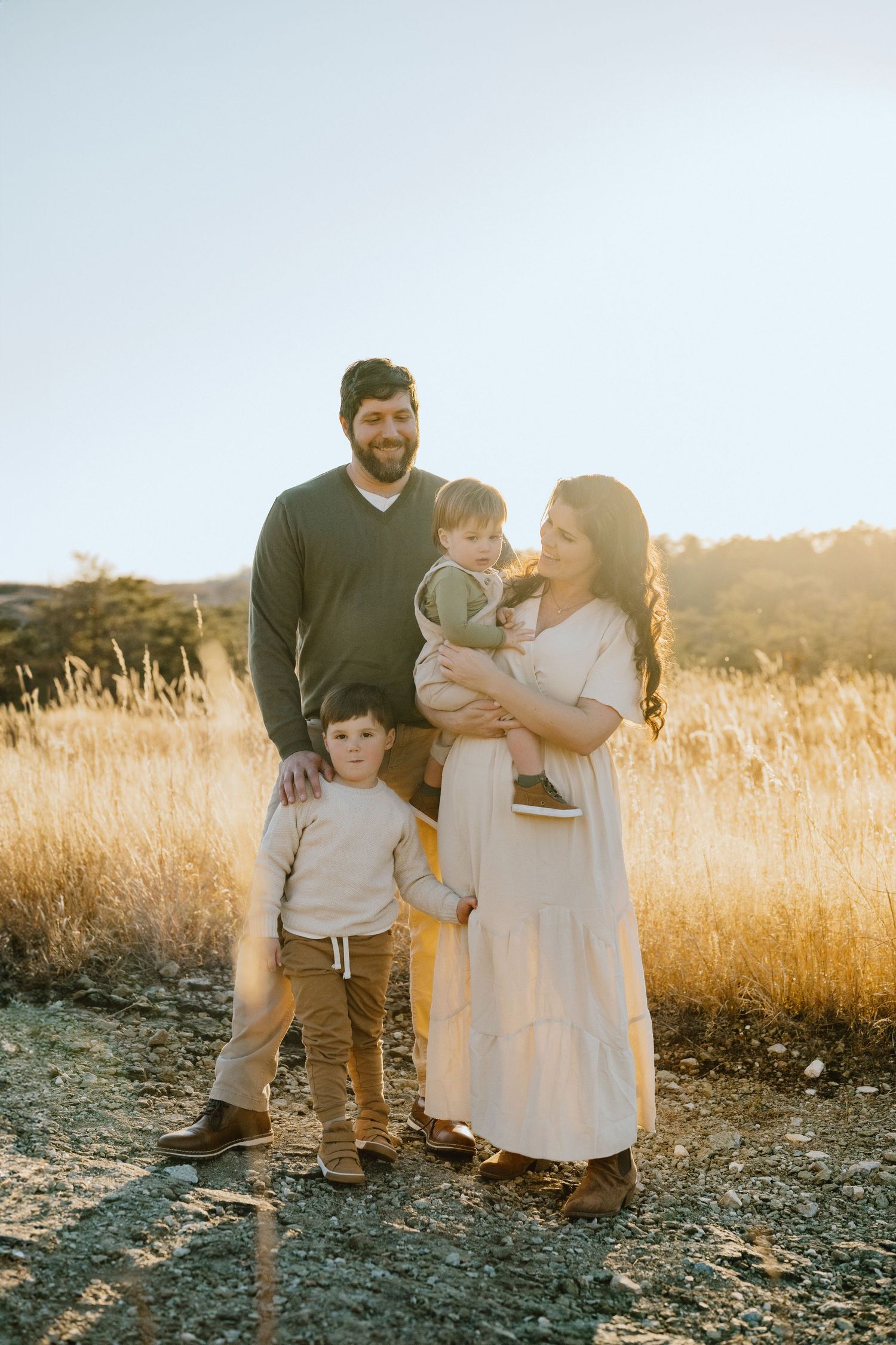 Family stands together in sunlit field with golden grass and mountain backdrop.