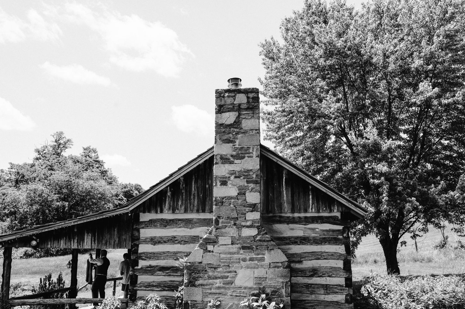 A rustic log cabin with stone chimney and wooden porch surrounded by trees.