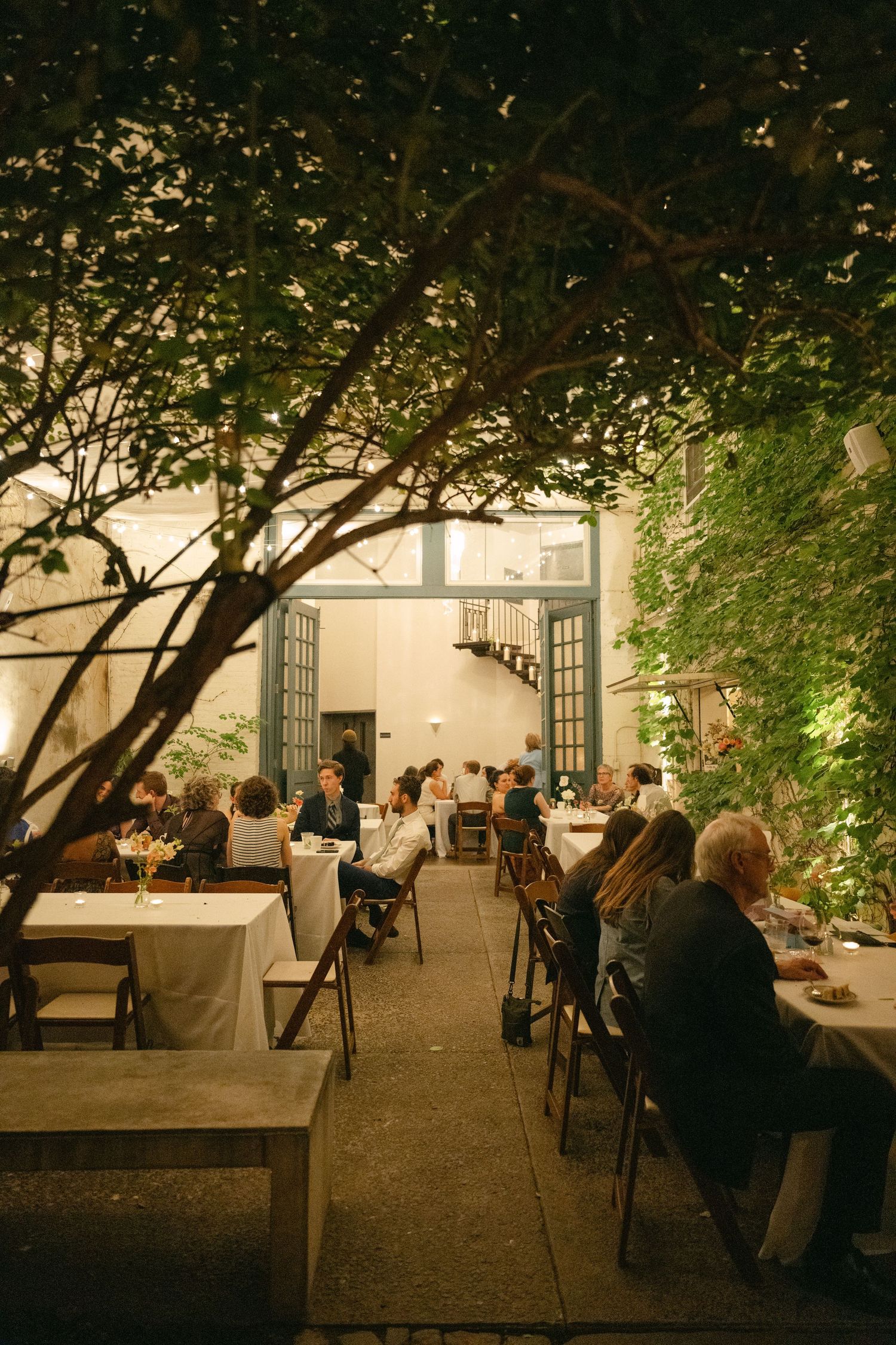 Outdoor restaurant seating area at dusk with tables arranged under trees and soft ambient lighting creating a cozy atmosphere.