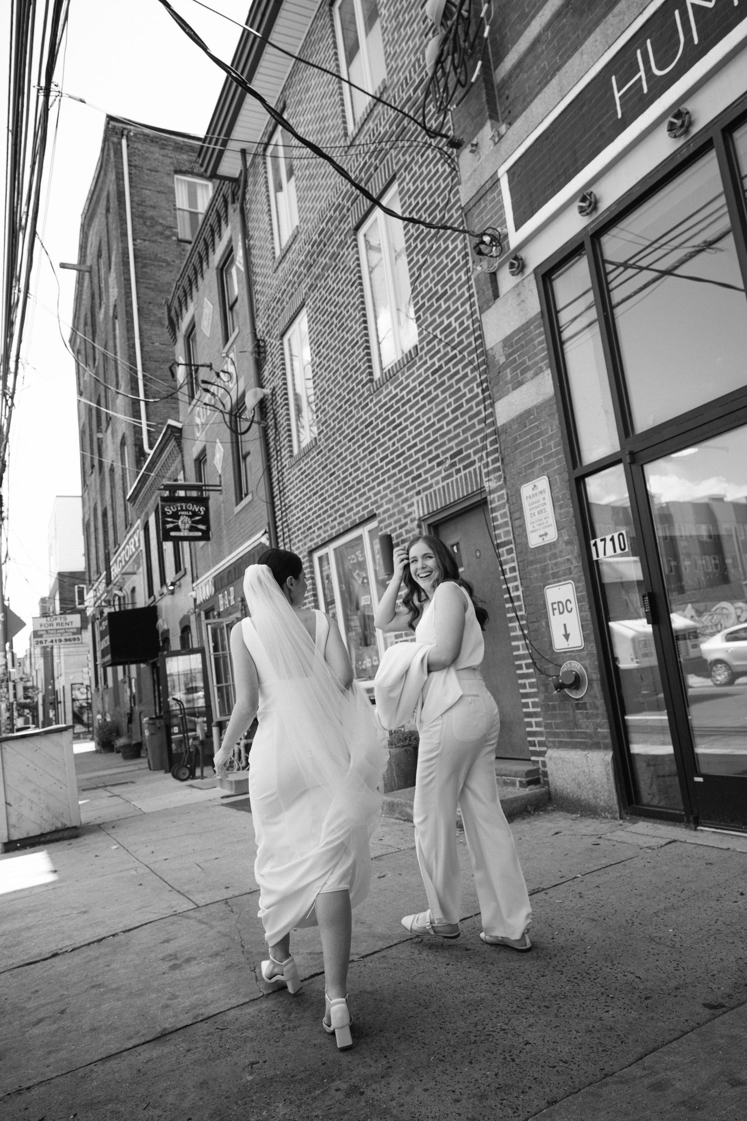 Figures in white dresses walk down an urban street lined with brick buildings.