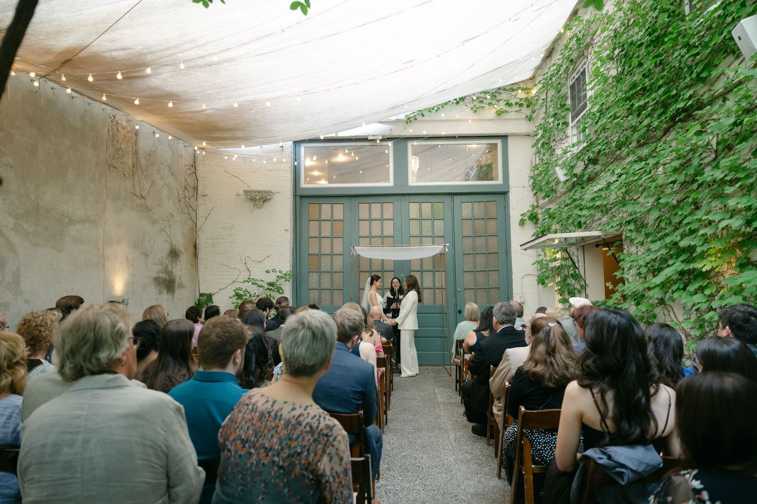 Outdoor wedding ceremony in a rustic courtyard with string lights and ivy-covered walls.