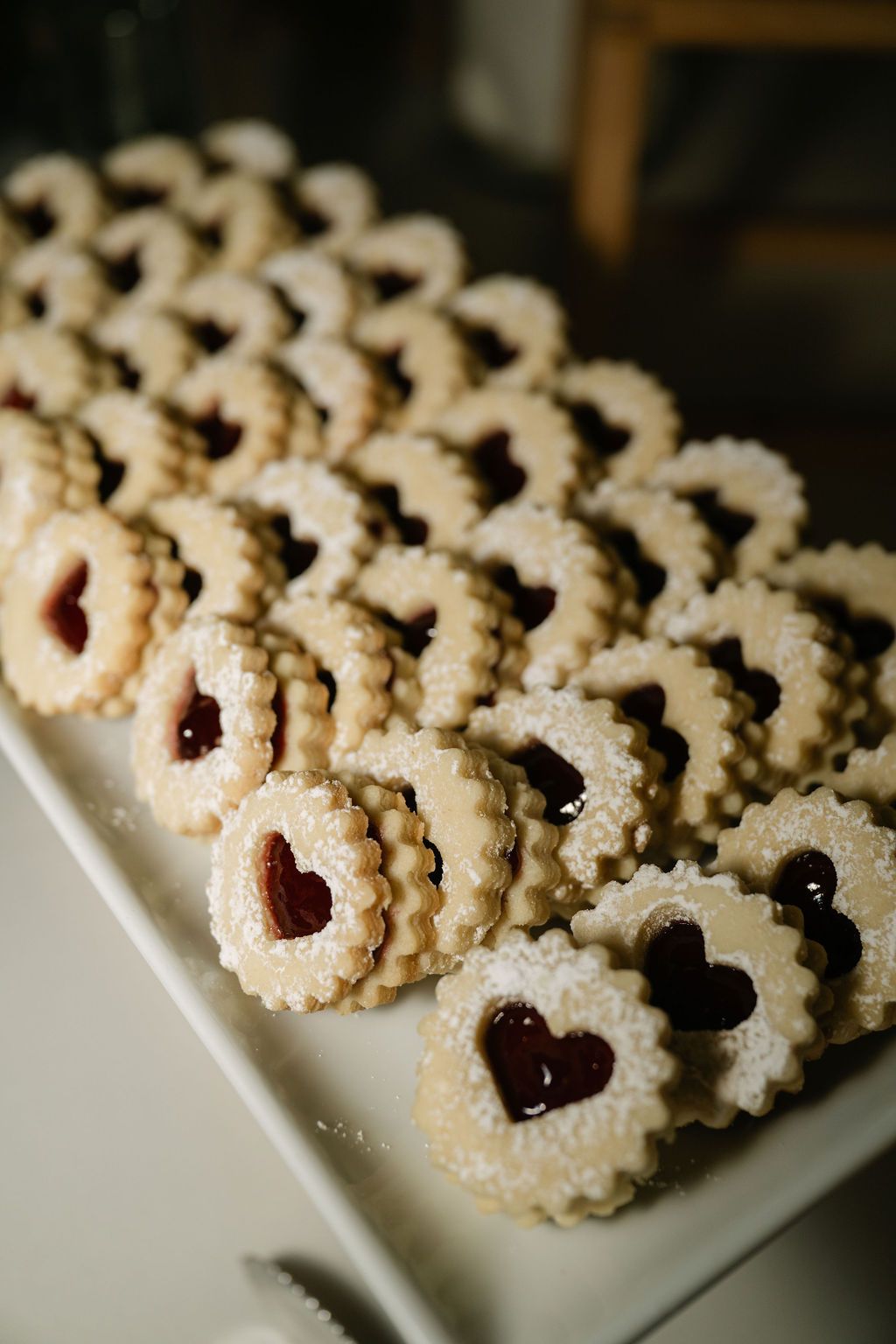 Rows of delicate heart-shaped linzer cookies with jam centers displayed on a white platter.