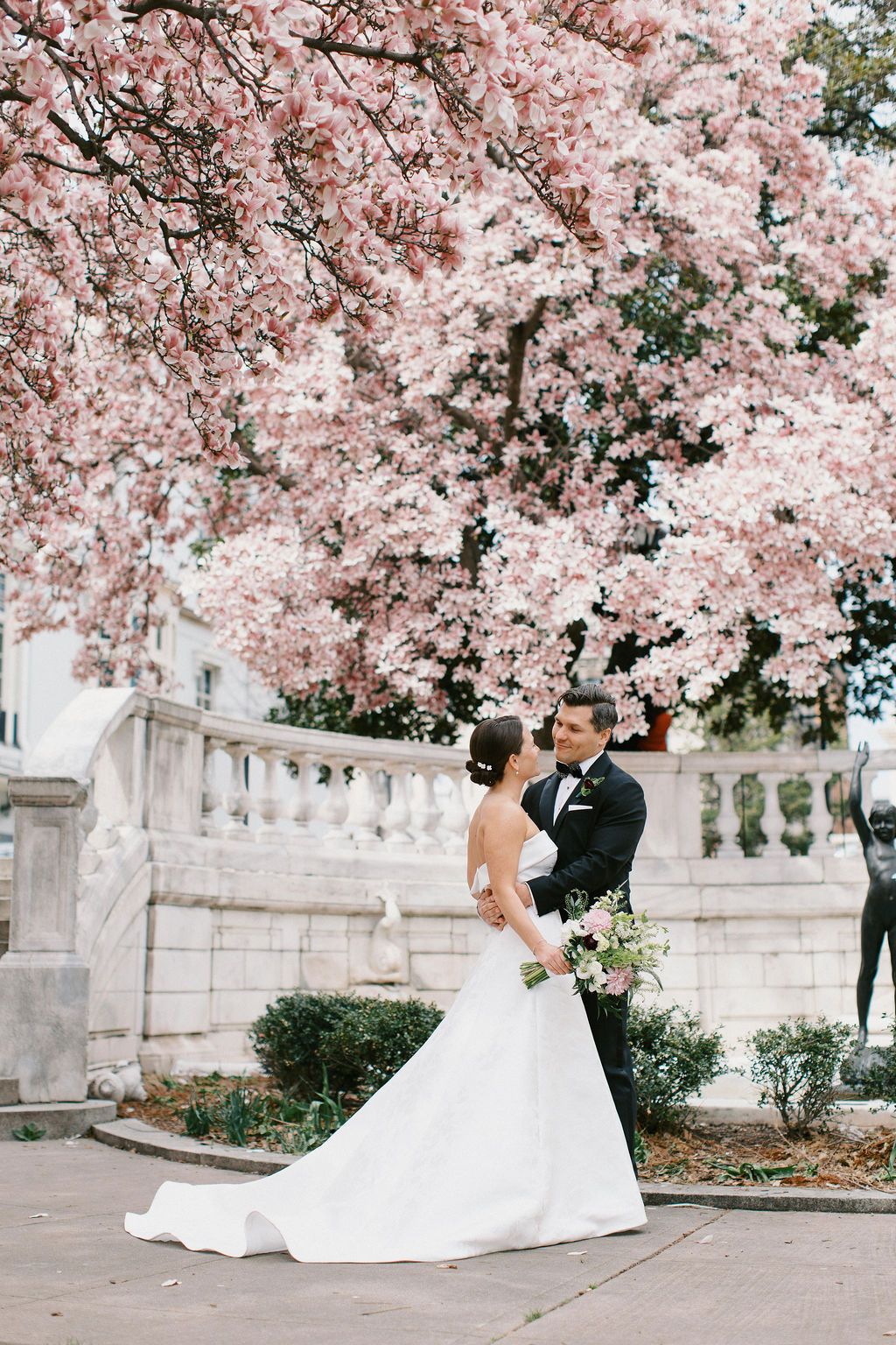 Elegant wedding couple poses under blooming cherry trees near stone balustrade.